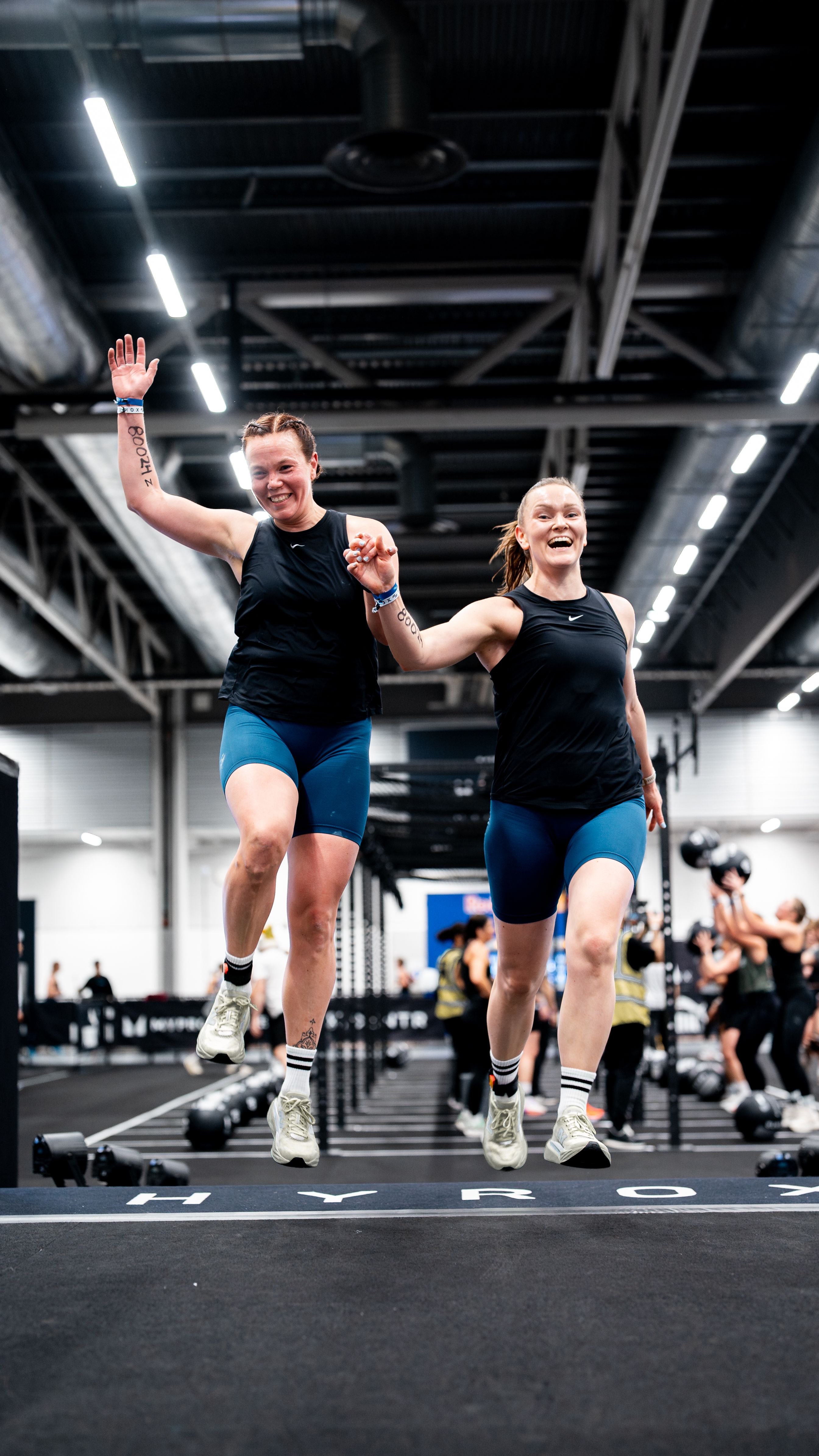 Two joyful women holding hands, mid-jump on a HYROX race mat.