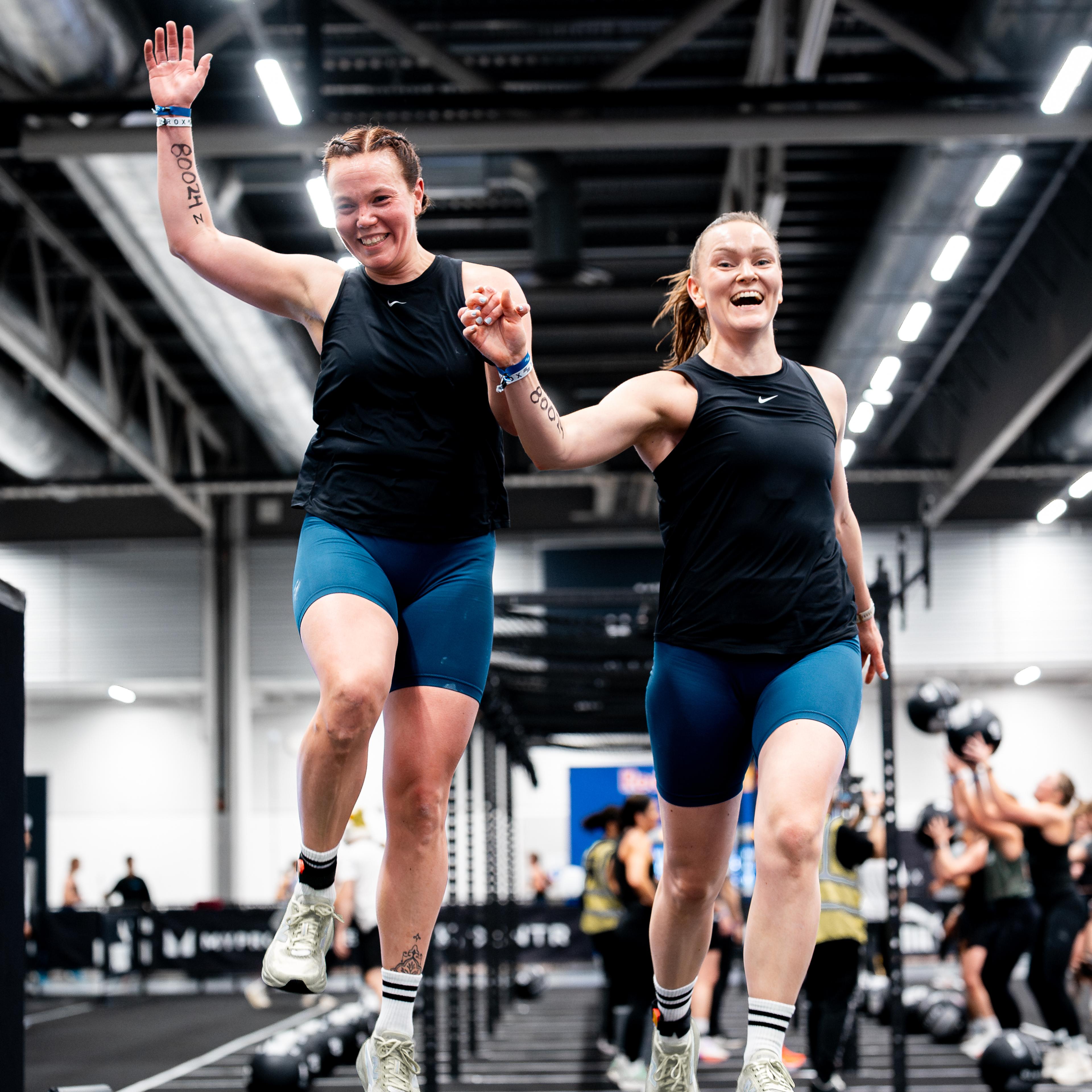 Two joyful women holding hands, mid-jump on a HYROX race mat.