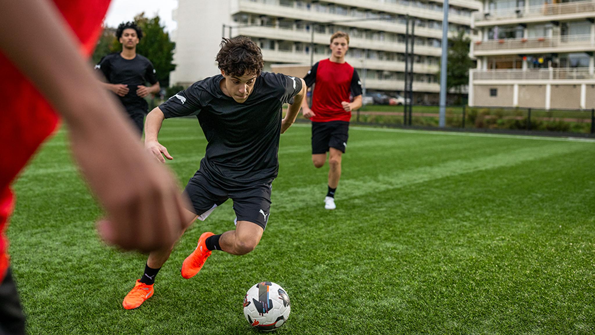 A young man in a black uniform dribbles a soccer ball on a green artificial turf field, with other players in the background.