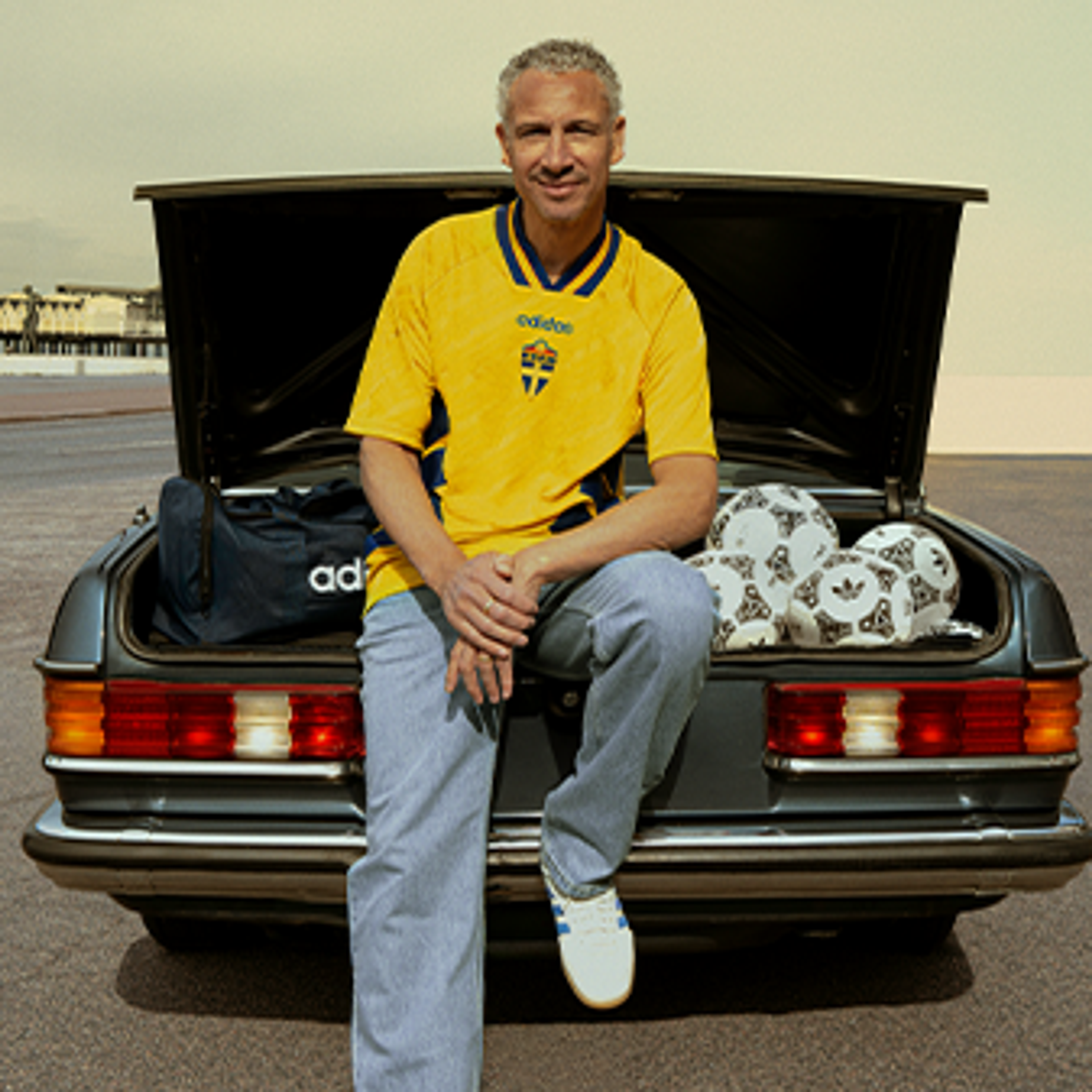 A man with salt-and-pepper hair sits on the open trunk of a dark car, wearing a yellow Adidas Sweden soccer jersey, jeans, and sneakers. An Adidas duffel bag and several Adidas soccer balls are in the trunk.