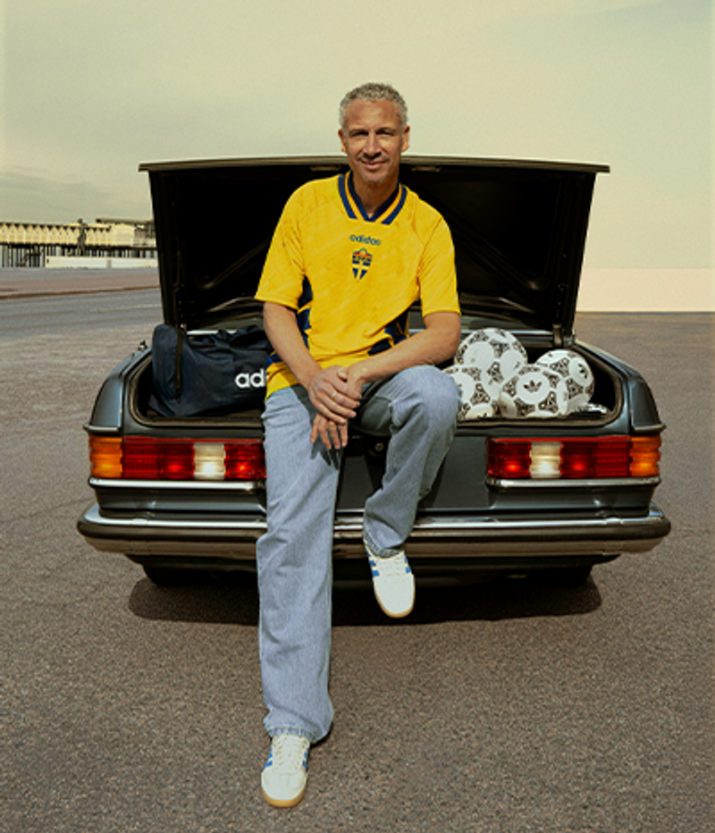A man in a yellow Sweden jersey sits on a car's open trunk with an Adidas bag and soccer balls.