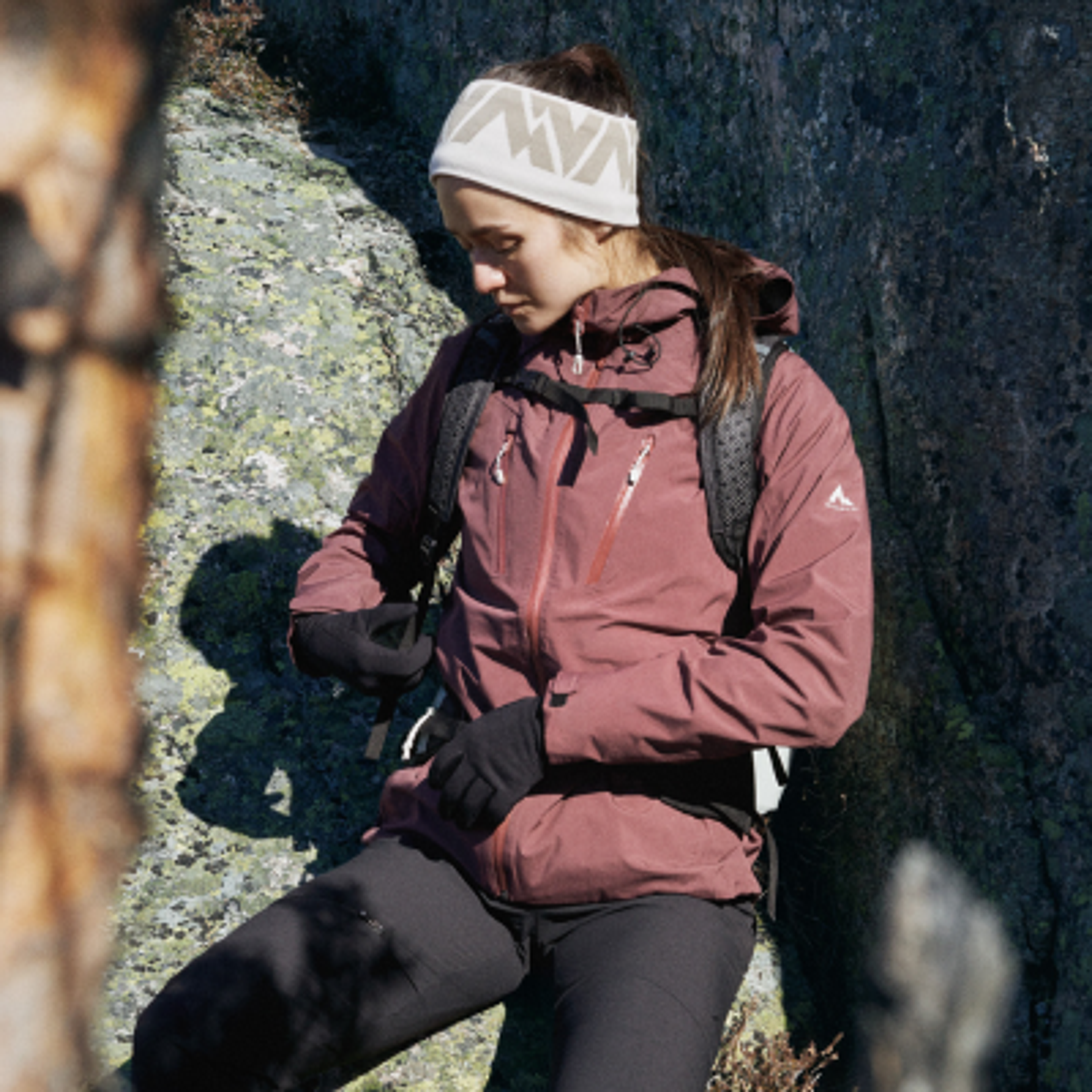 A woman in a maroon hiking jacket, white headband, and backpack adjusts her gear in a rocky outdoor setting.