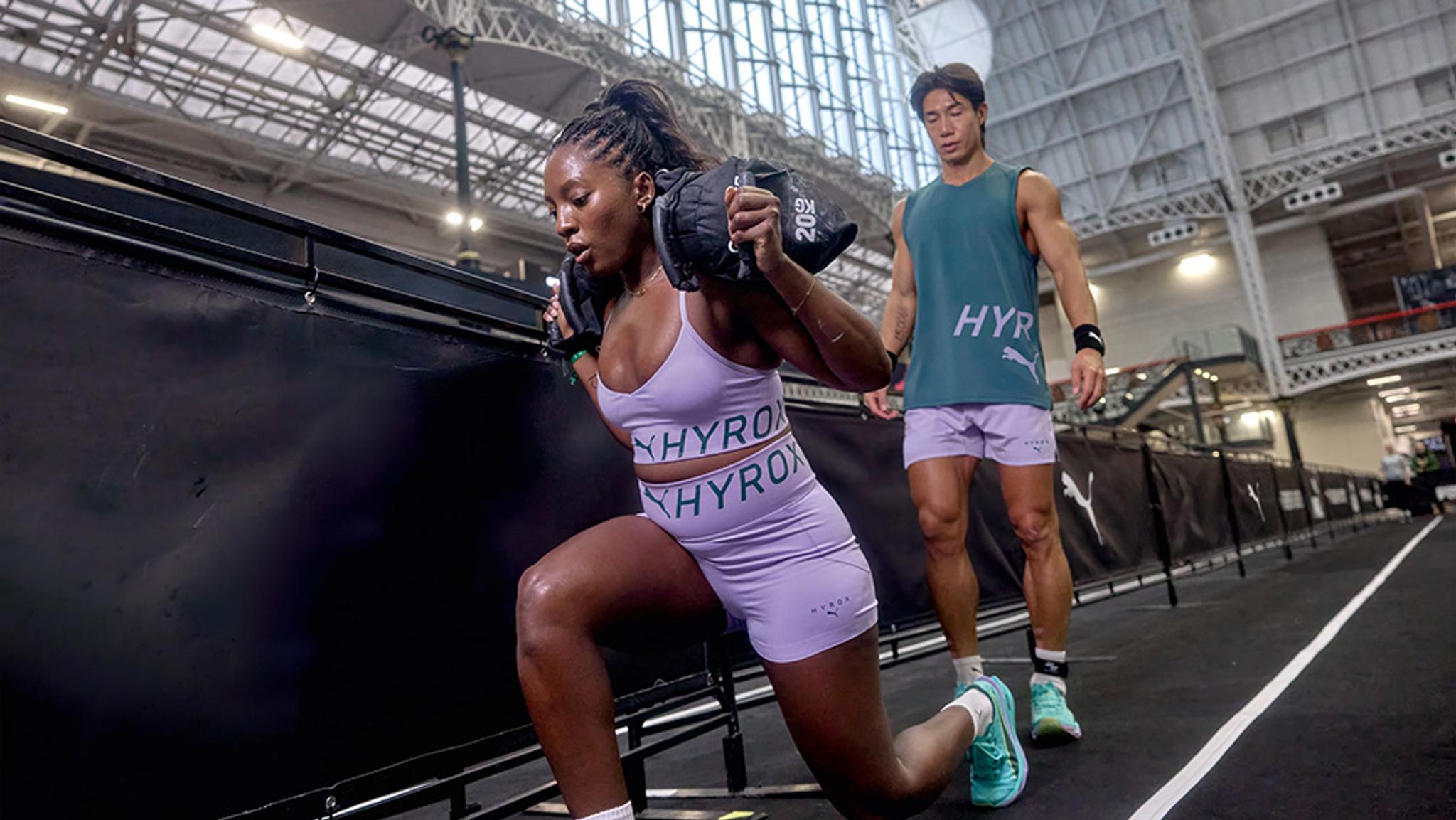 A woman in HYROX activewear lunges with a sandbag while a man watches in a gym.
