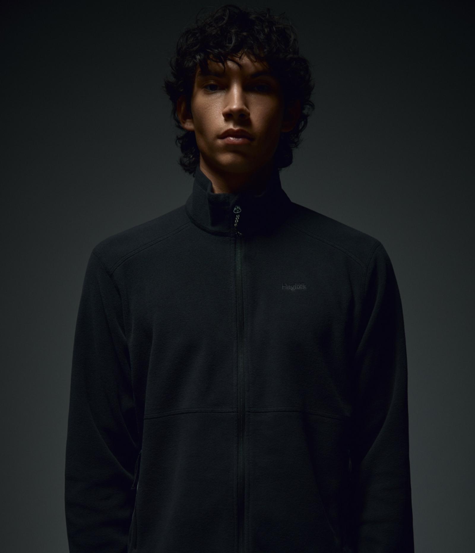 A young man with dark, curly hair wears a black zippered fleece jacket and looks directly ahead.