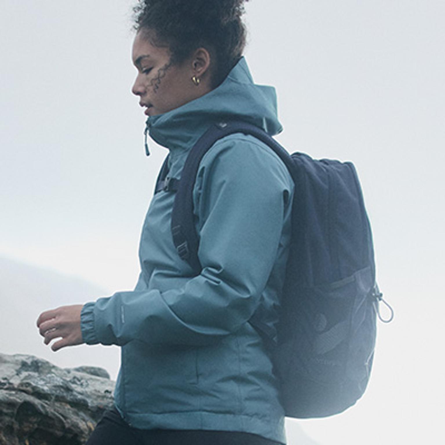 a woman wearing a blue jacket and a black backpack is walking on a rock .
