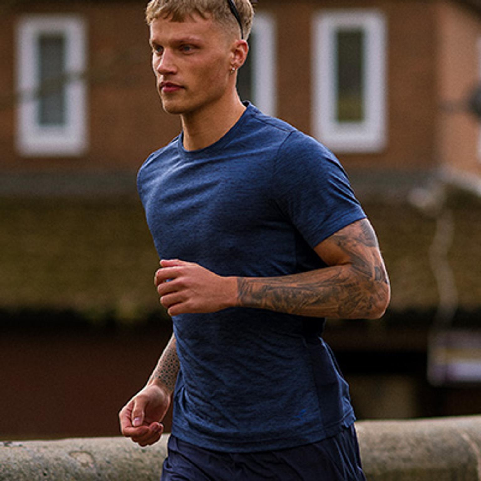 A young man in a blue t-shirt with tattooed arms runs outdoors.