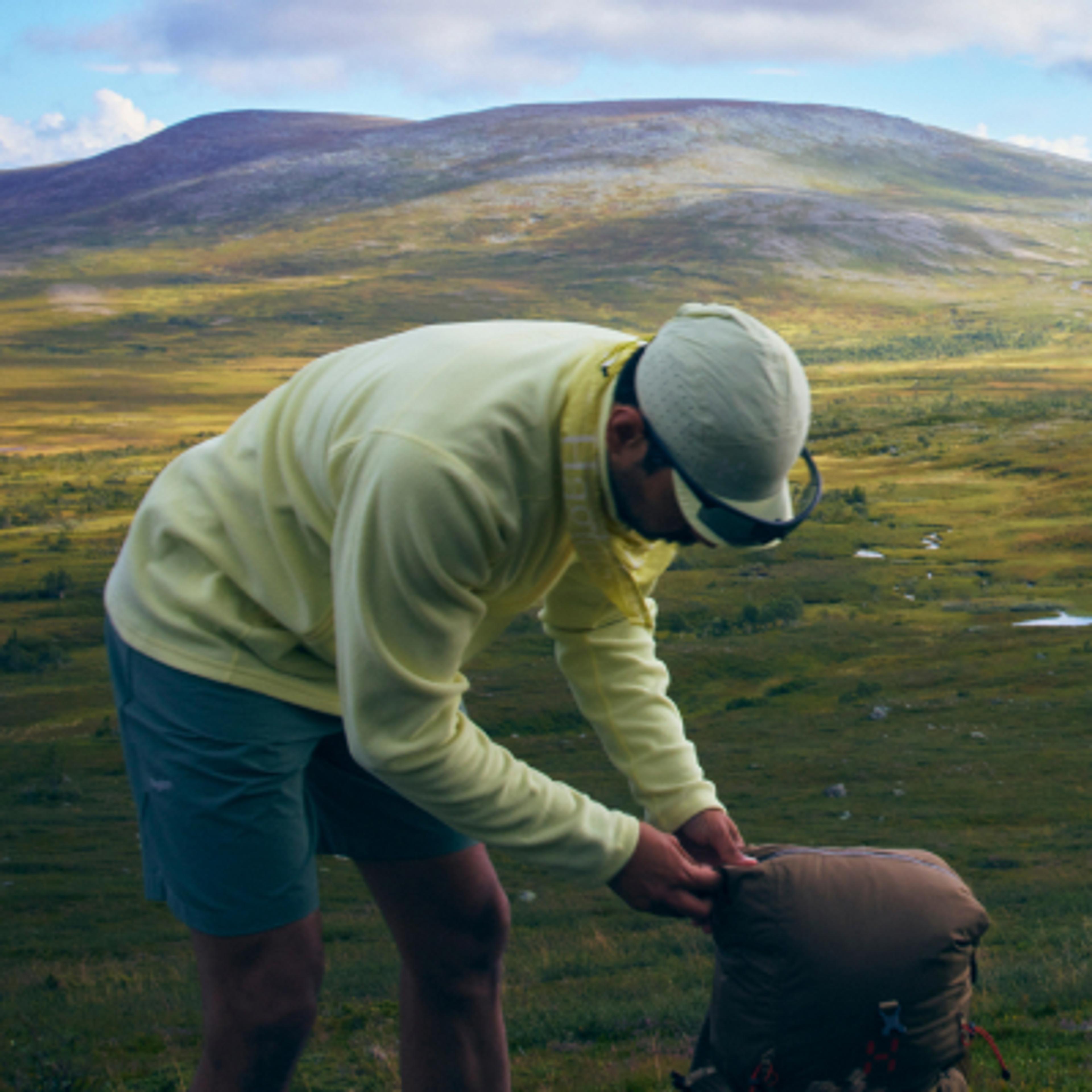 A hiker in a yellow shirt and cap bends over to adjust his backpack in a wide, grassy mountain valley.