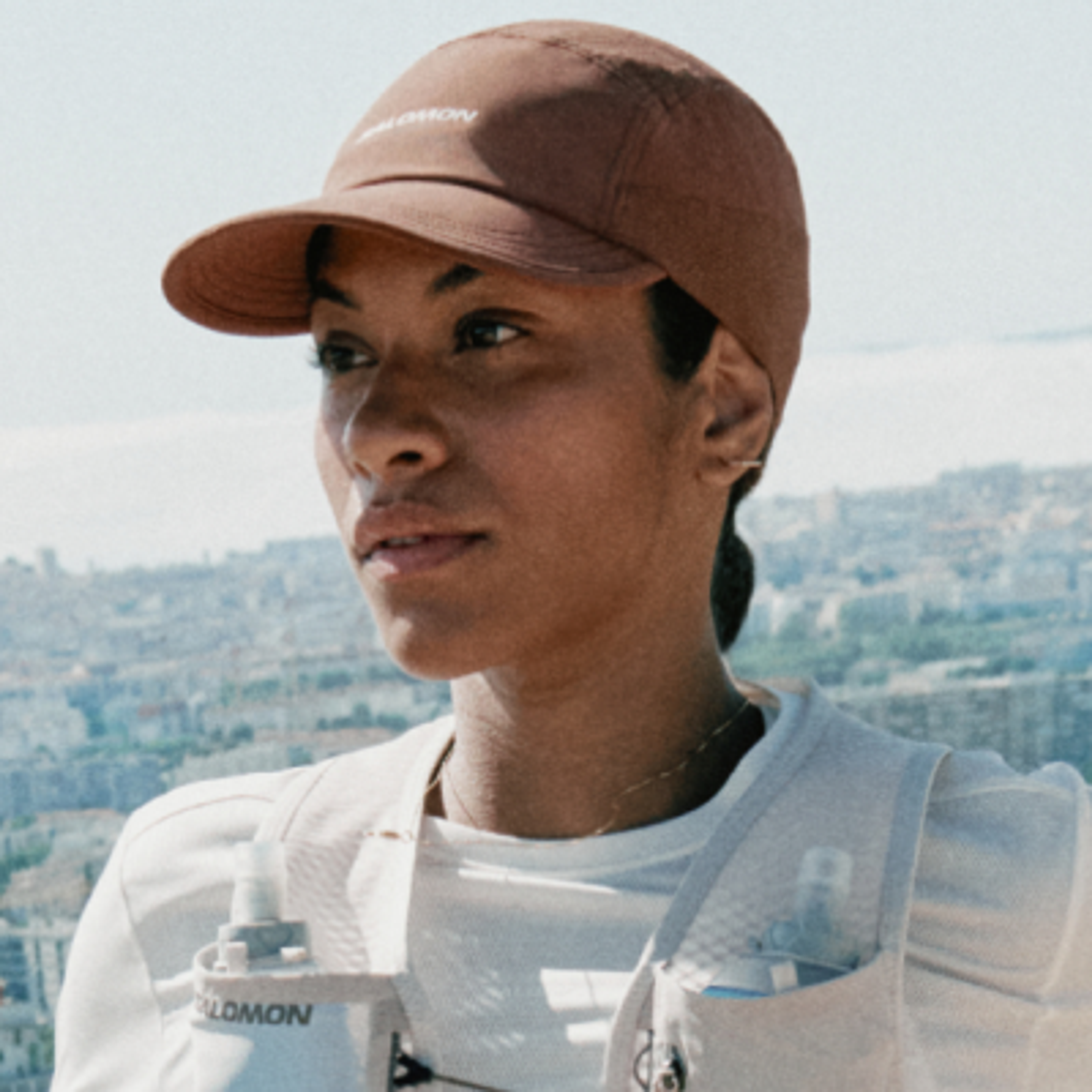 A woman in a Salomon cap and hydration vest looks towards a city view.