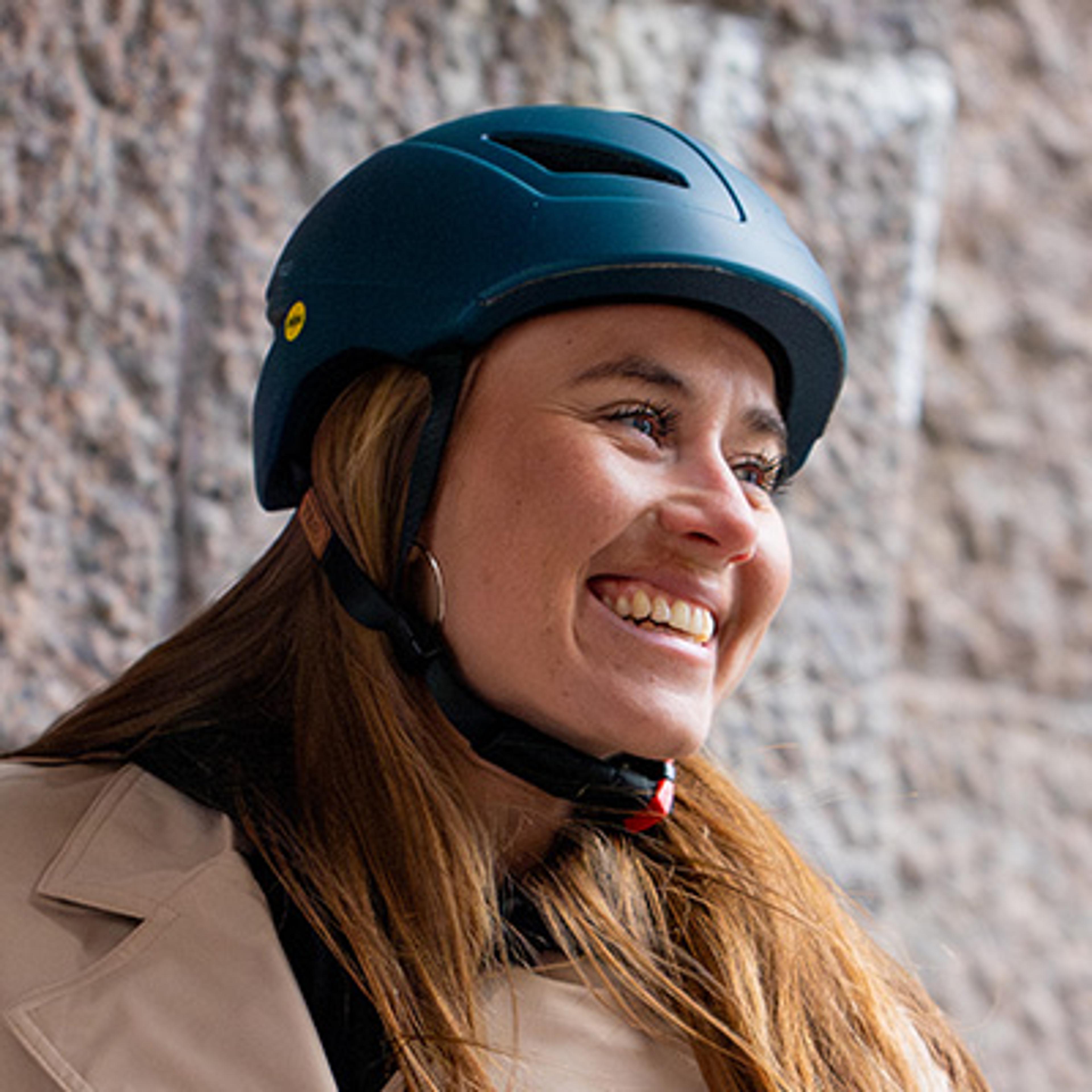 A smiling woman wearing a dark teal helmet and a tan coat, looking to her right.