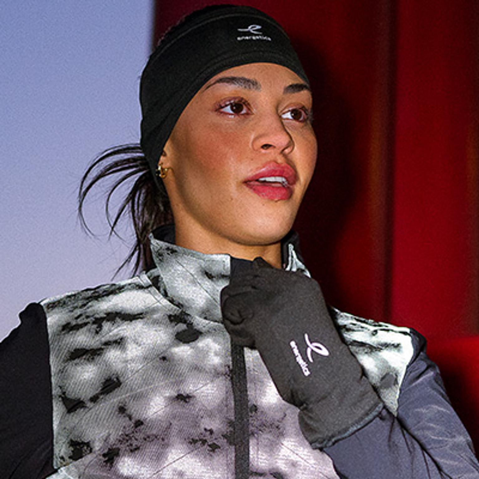 A woman wearing an Energetics headband, grey and black patterned vest, and gloves adjusts her collar while looking up.