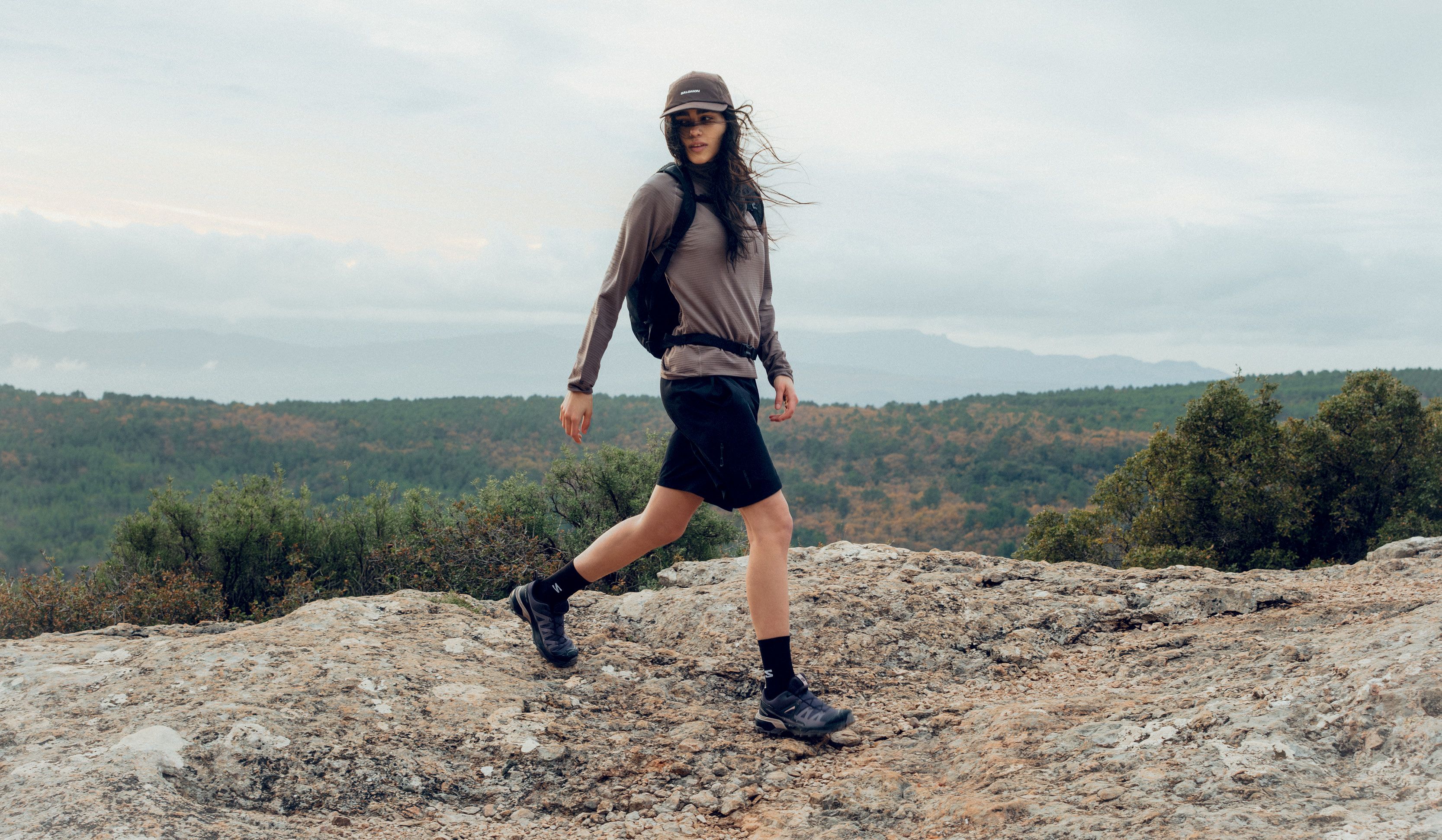 A woman in activewear and a cap mid-stride on a rocky trail above a forested landscape.