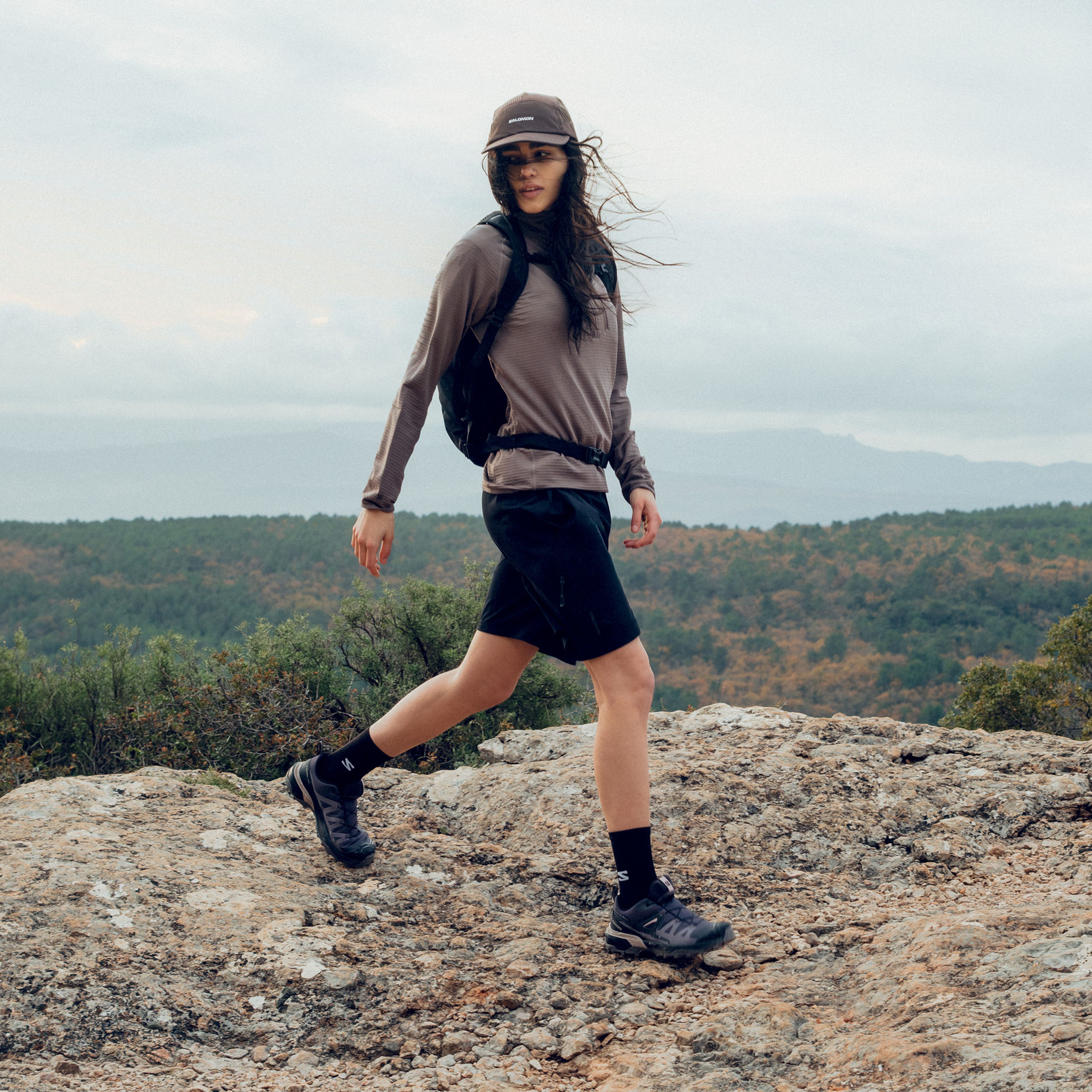 A woman in activewear and a cap mid-stride on a rocky trail above a forested landscape.