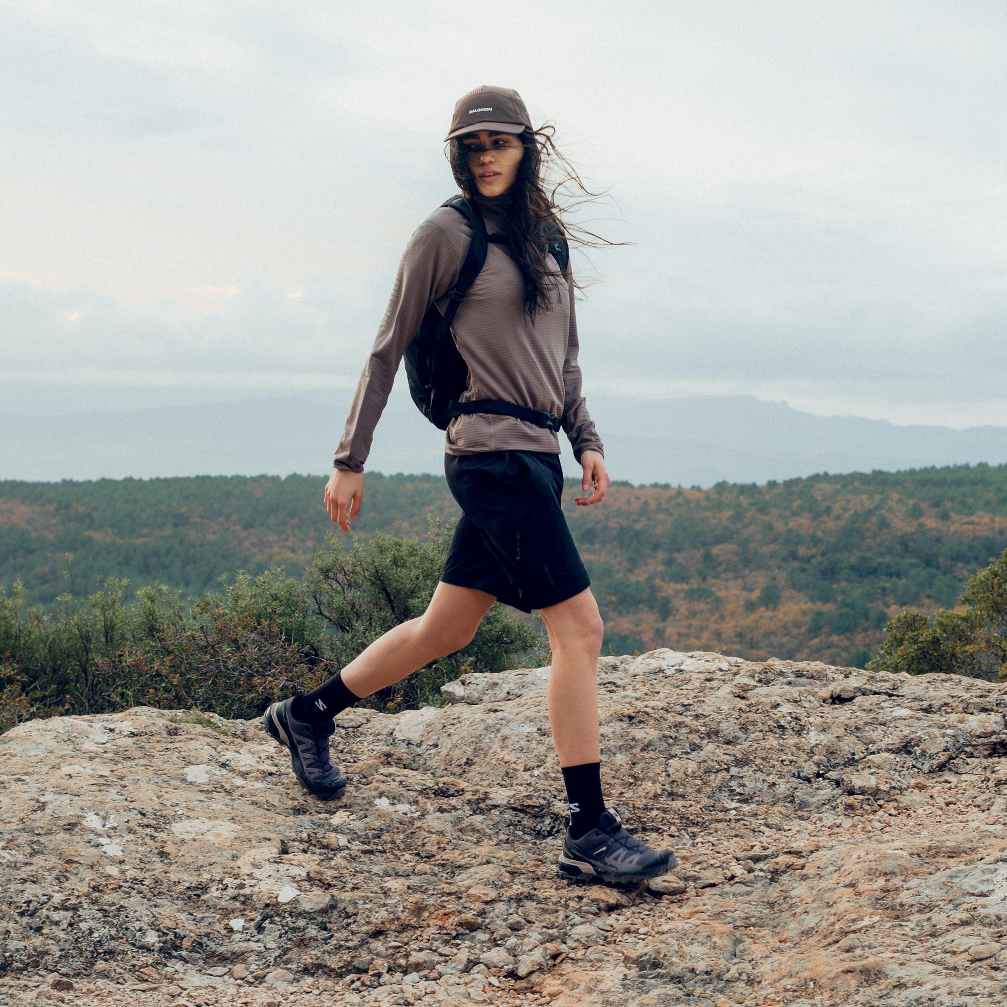 A woman in activewear and a cap mid-stride on a rocky trail above a forested landscape.