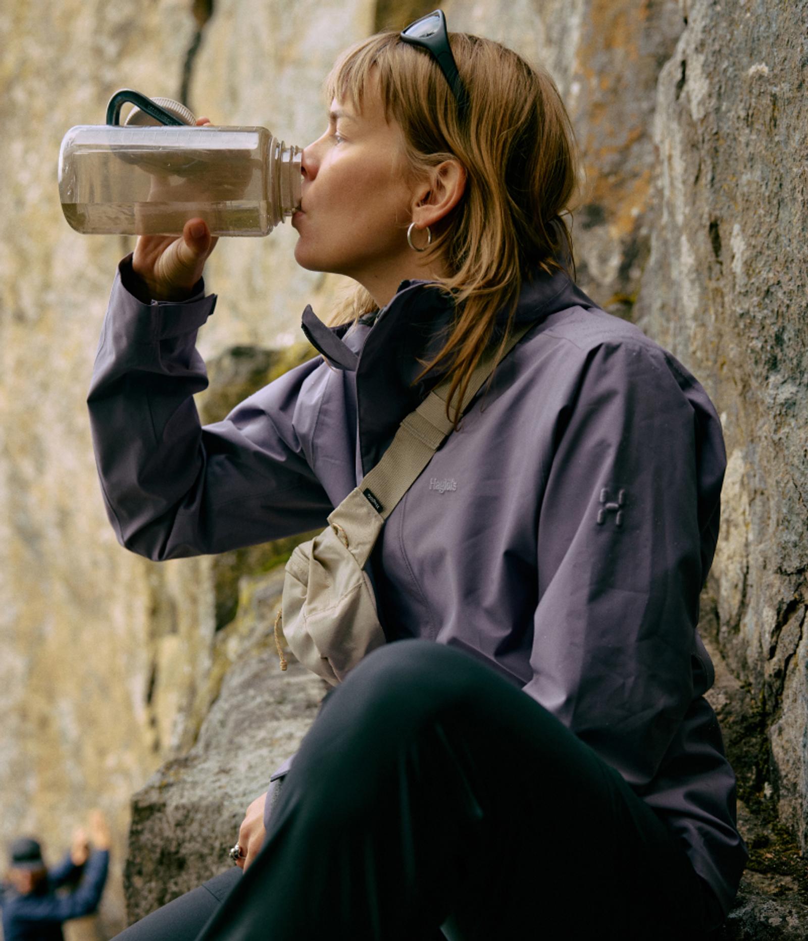 A woman in outdoor gear drinks from a water bottle against a rocky background.