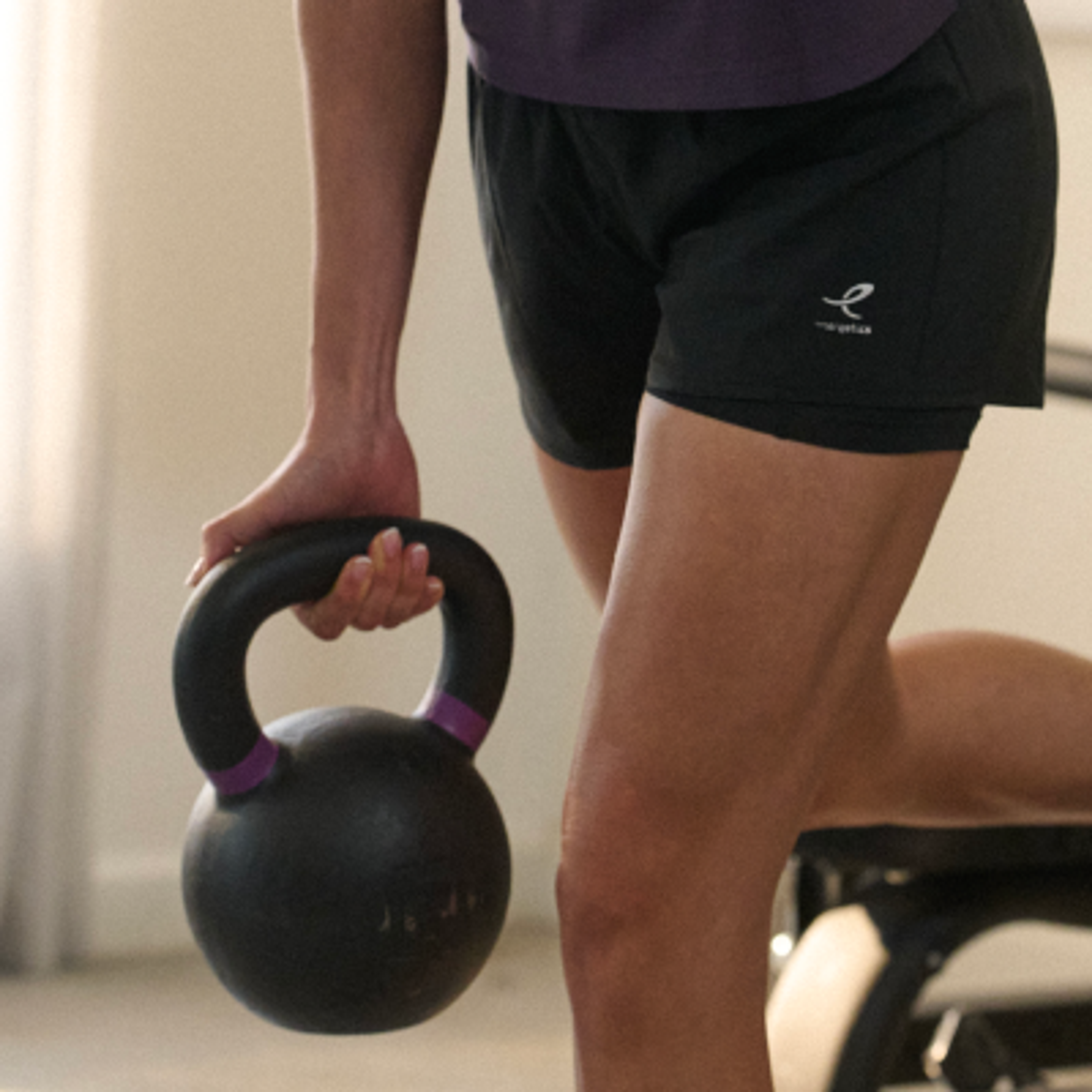 A person's hand grips a black kettlebell with purple stripes, wearing black athletic shorts.