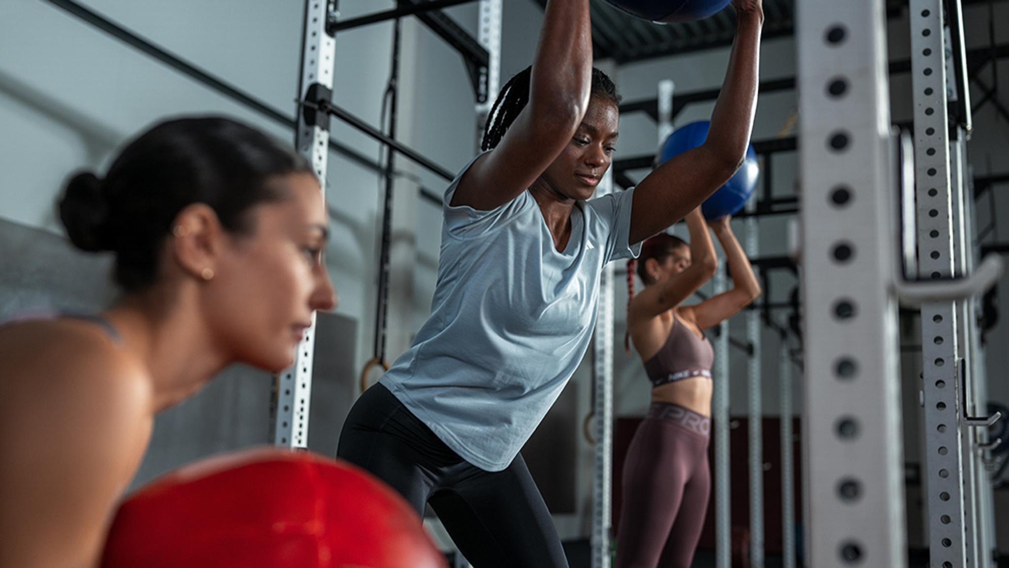 Three women exercise with medicine balls in a gym, two holding balls overhead.