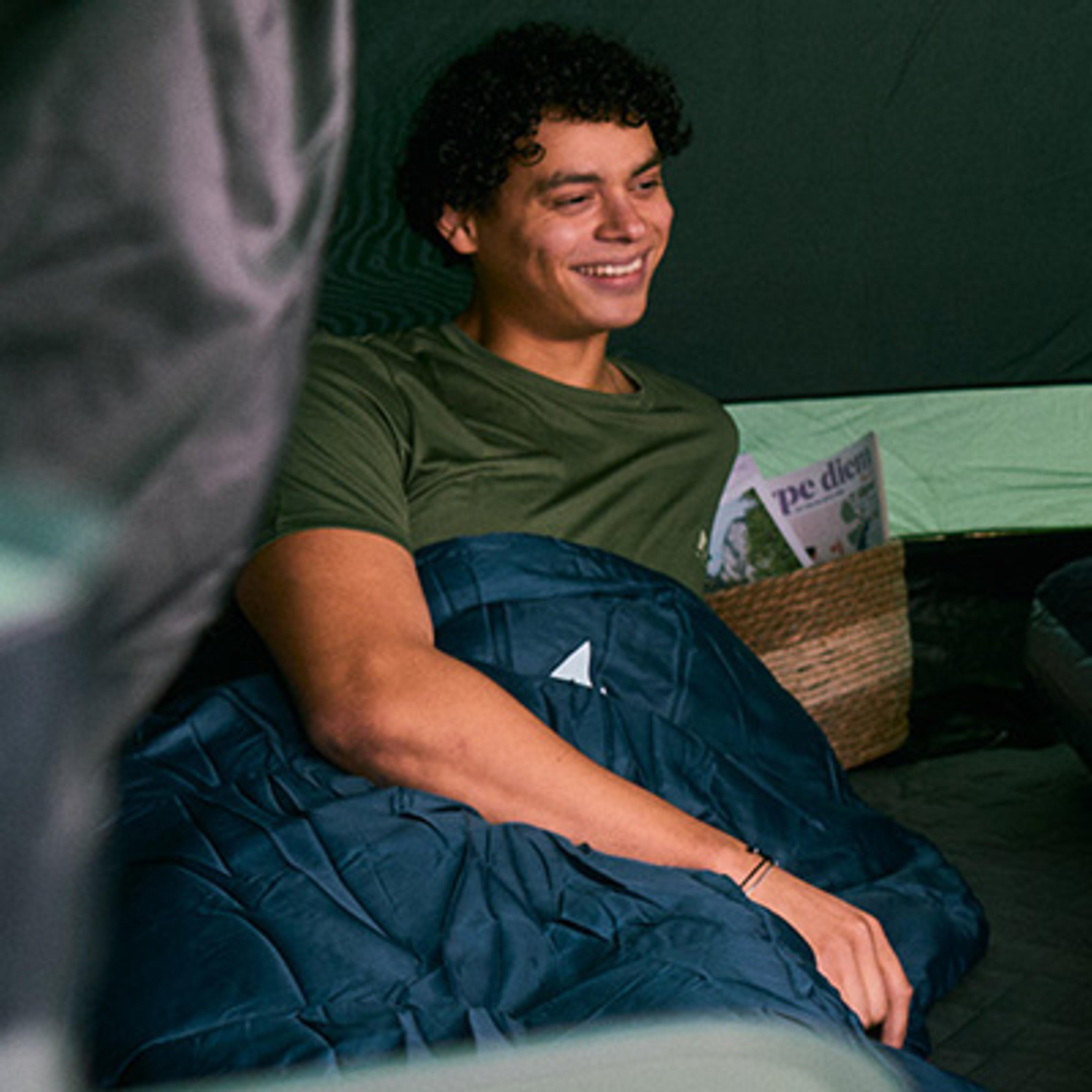 A smiling person sits inside a tent, covered by a blue blanket.