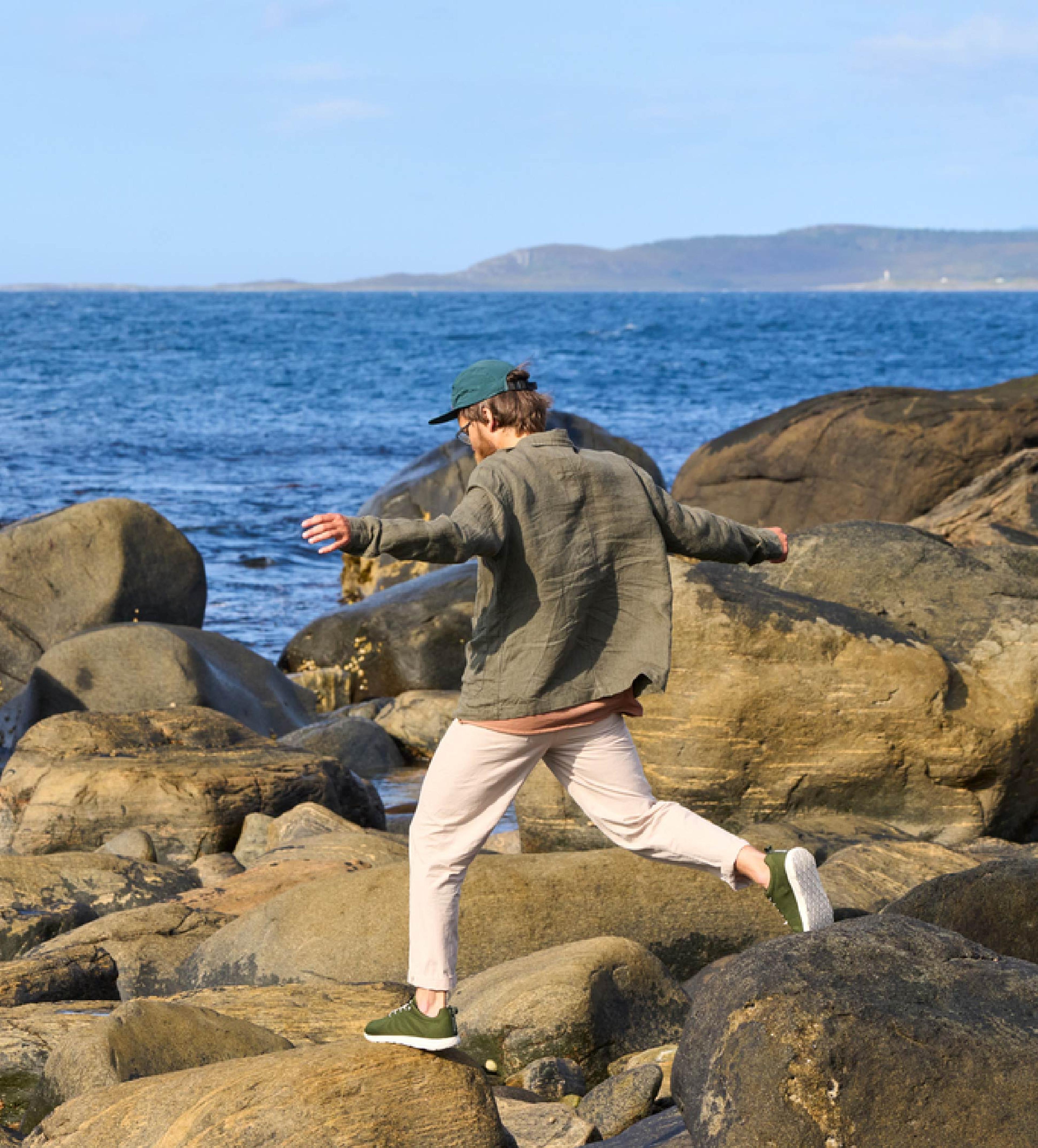 A person in a green jacket and cap steps across rocks by the ocean.