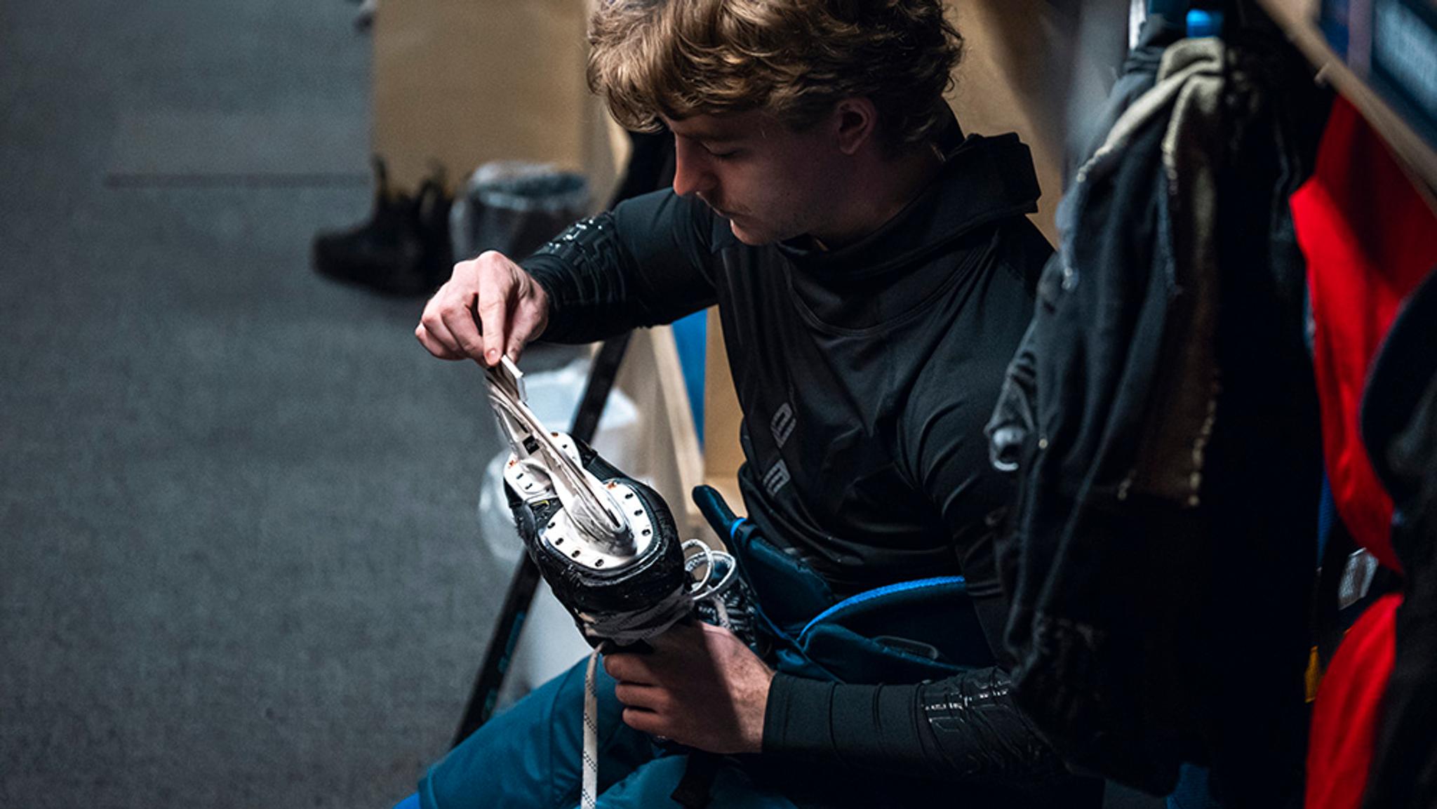 A young man sitting on a bench and lacing up an ice skate.