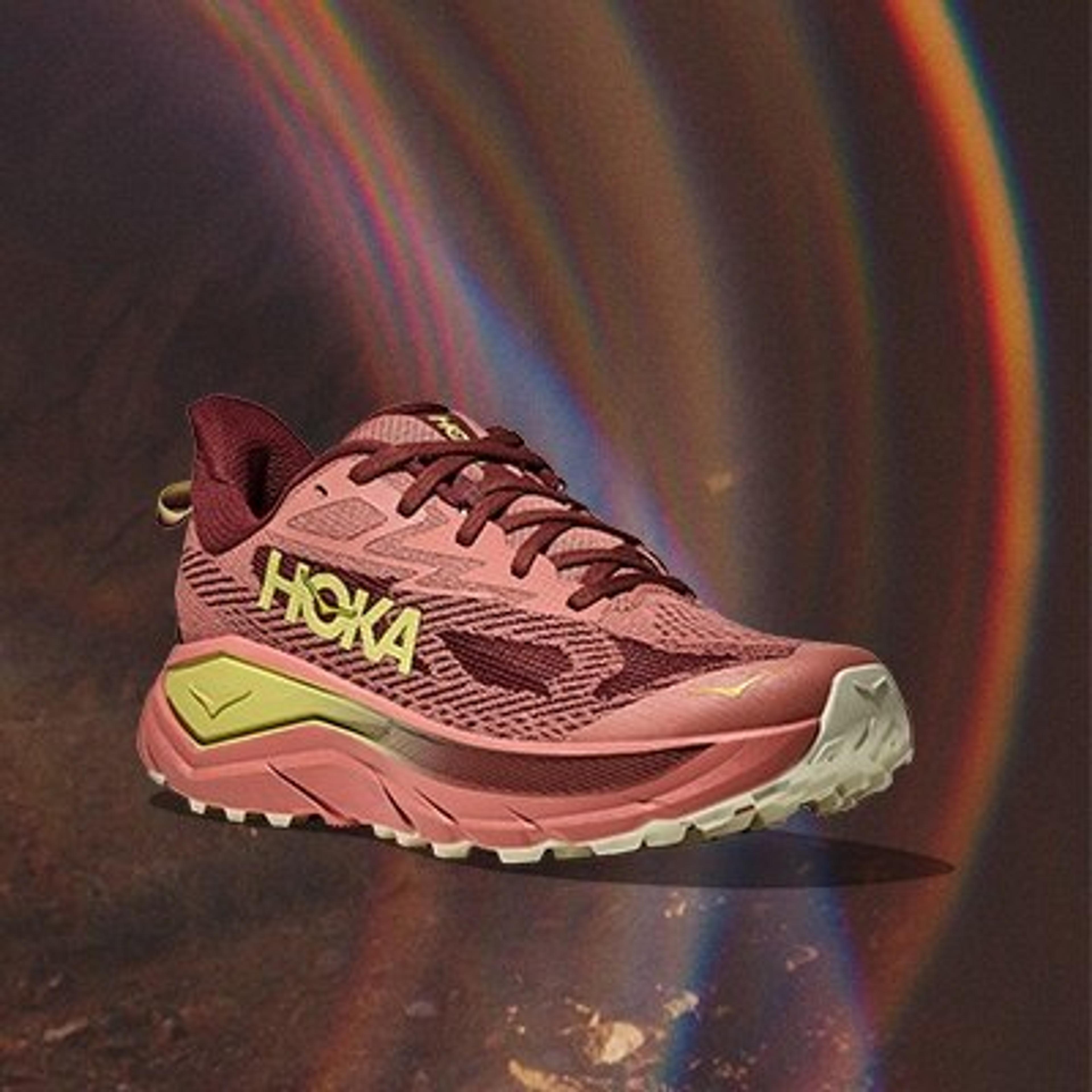 a pair of pink hoka one one trail running shoes are sitting on top of a dirt surface with a rainbow in the background .