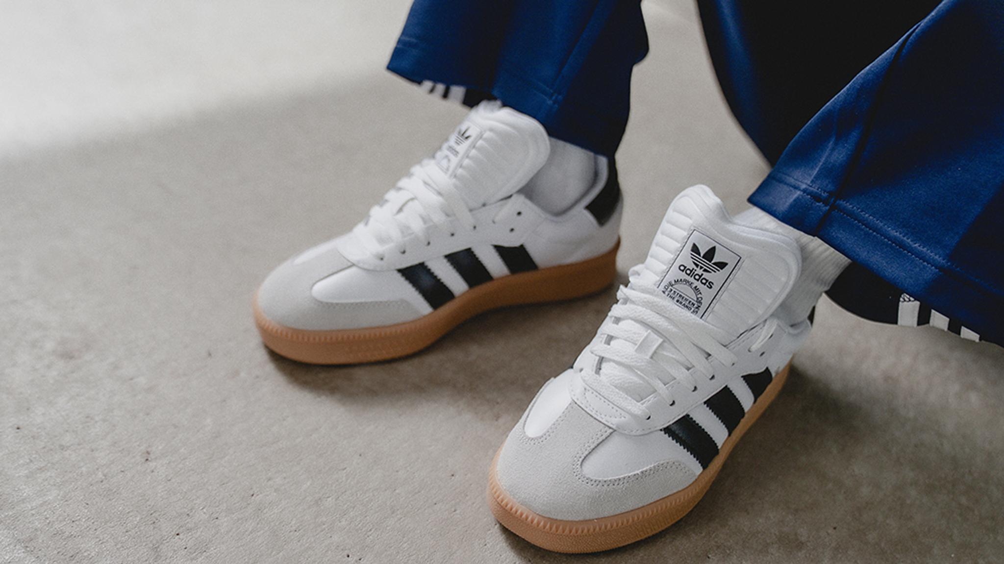 A person's feet wearing white Adidas Samba sneakers with black stripes and gum soles, on a concrete floor.