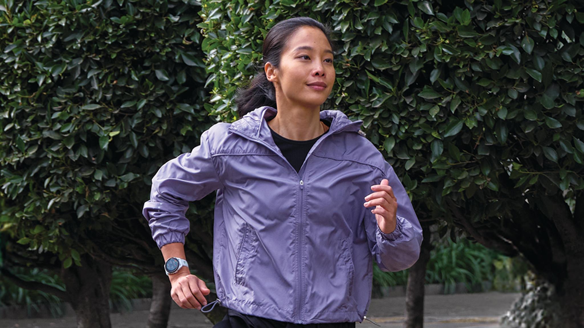 A woman in a purple jacket and smartwatch jogs outdoors.