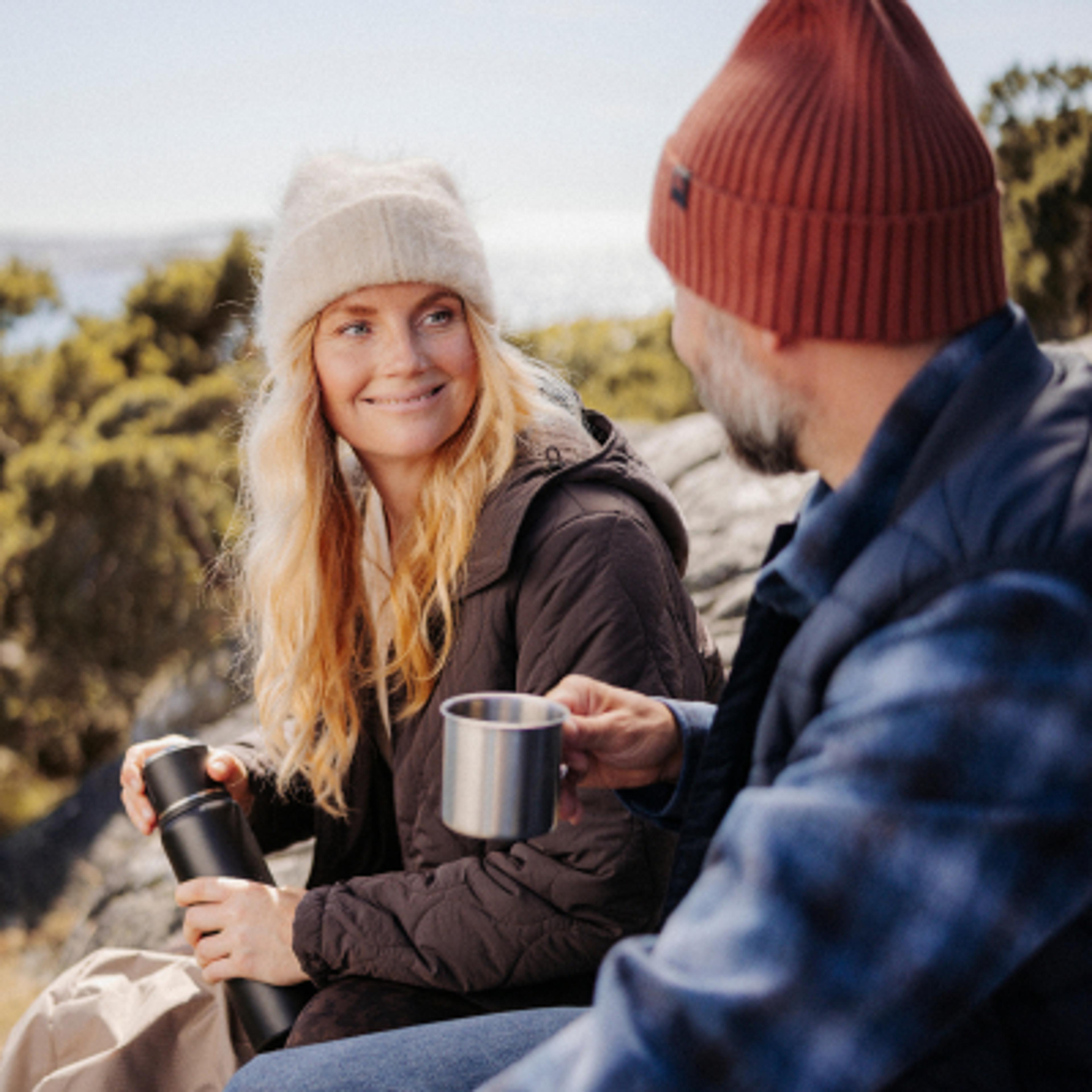 A smiling woman holding a thermos looks at a man holding a mug while sitting outdoors.