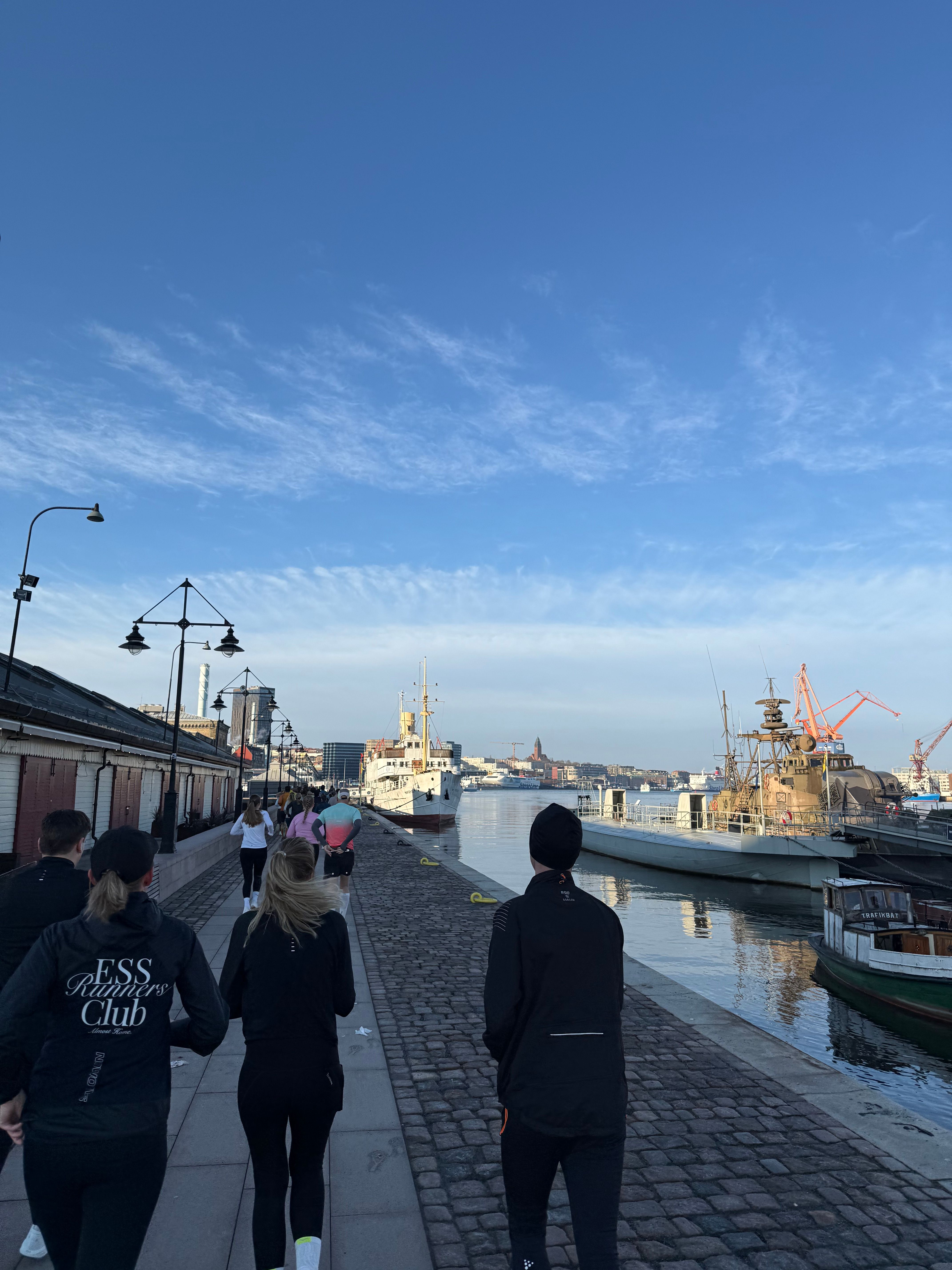 People running along a cobblestone quay with boats and a city in the background under a blue sky.