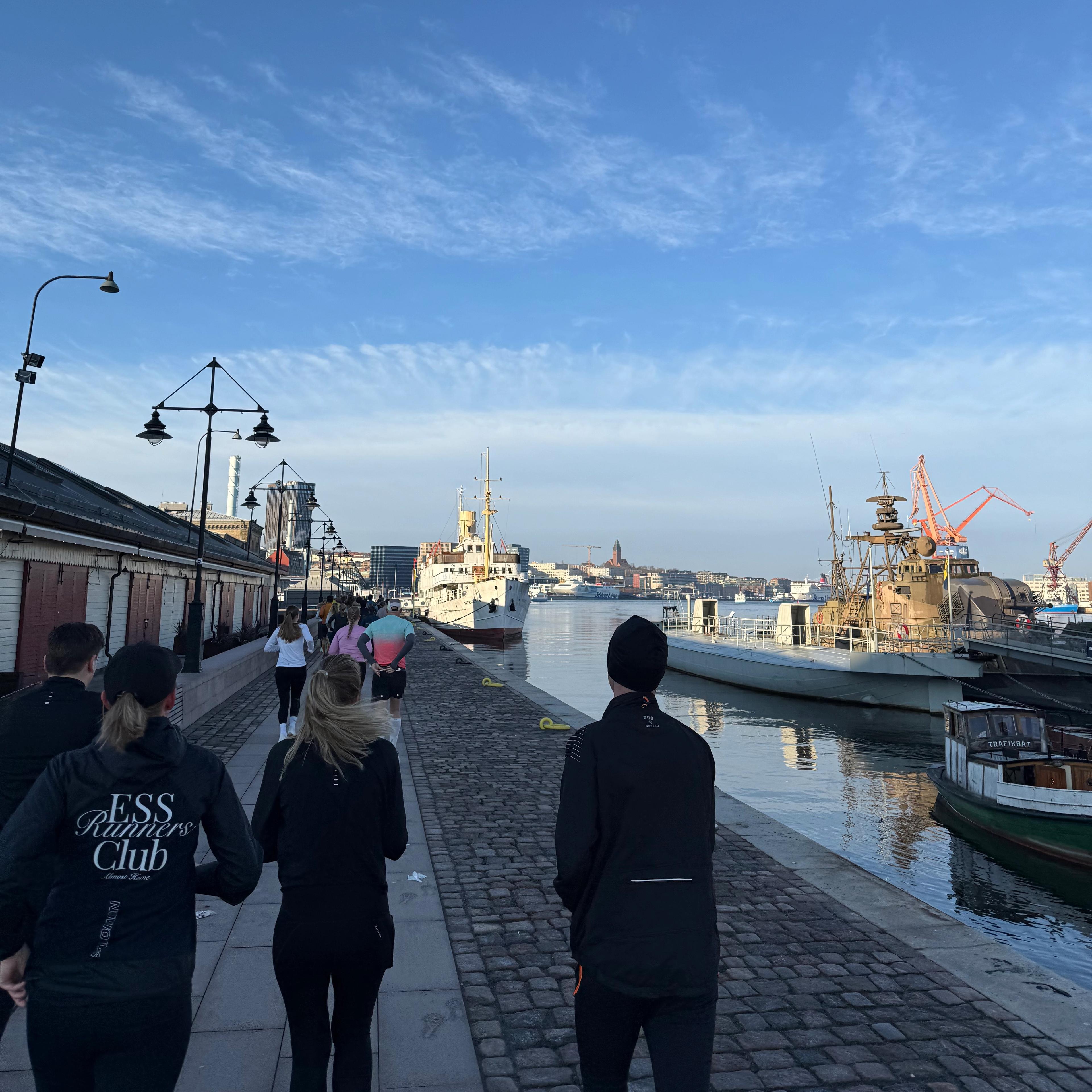 People running along a cobblestone quay with boats and a city in the background under a blue sky.