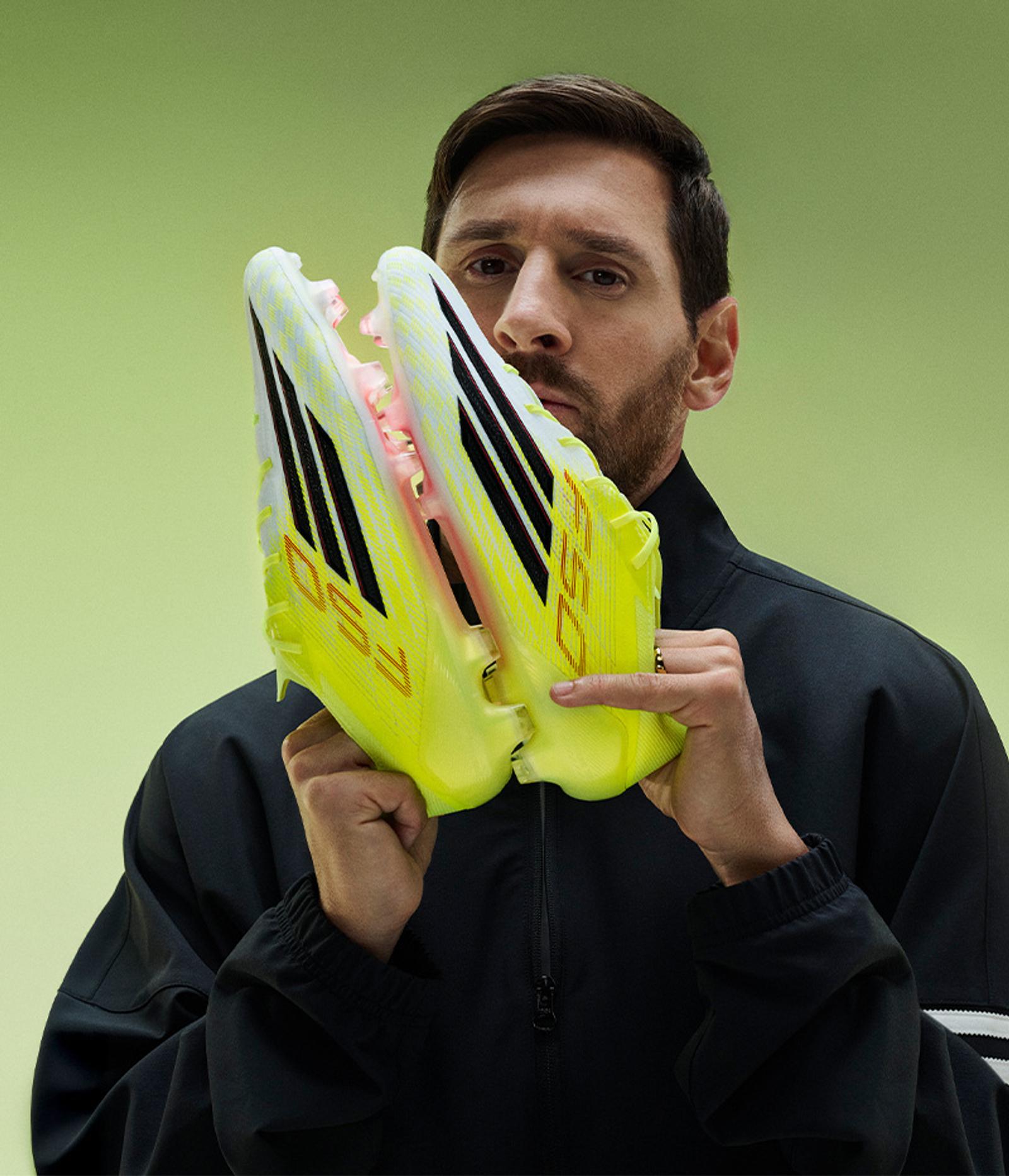 Lionel Messi holding neon yellow soccer cleats in front of his face against a light green background.