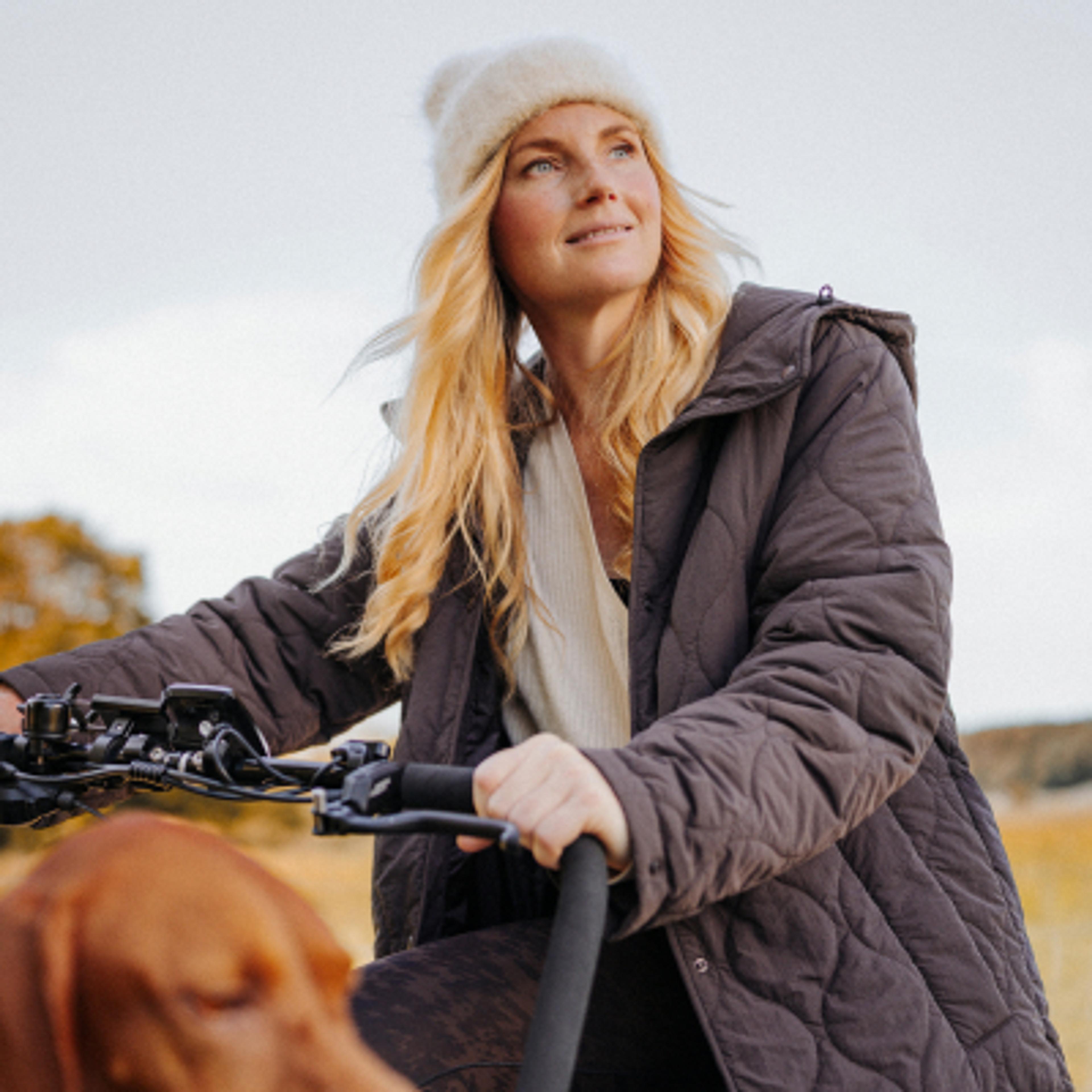A blonde woman in a white beanie and quilted jacket rides a bike, smiling, with a brown dog in the foreground.