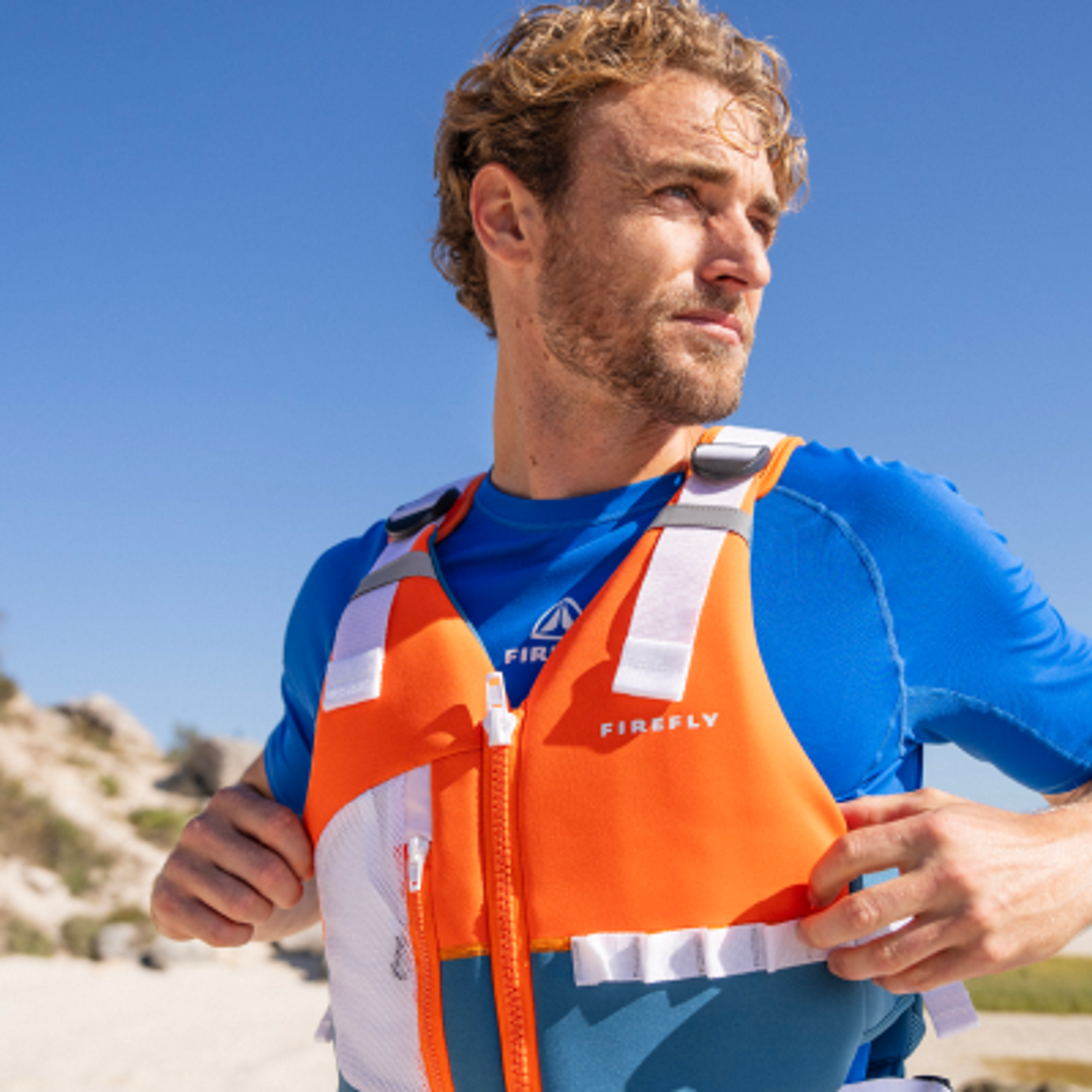 A man adjusting an orange and blue Firefly life vest on a sunny beach.