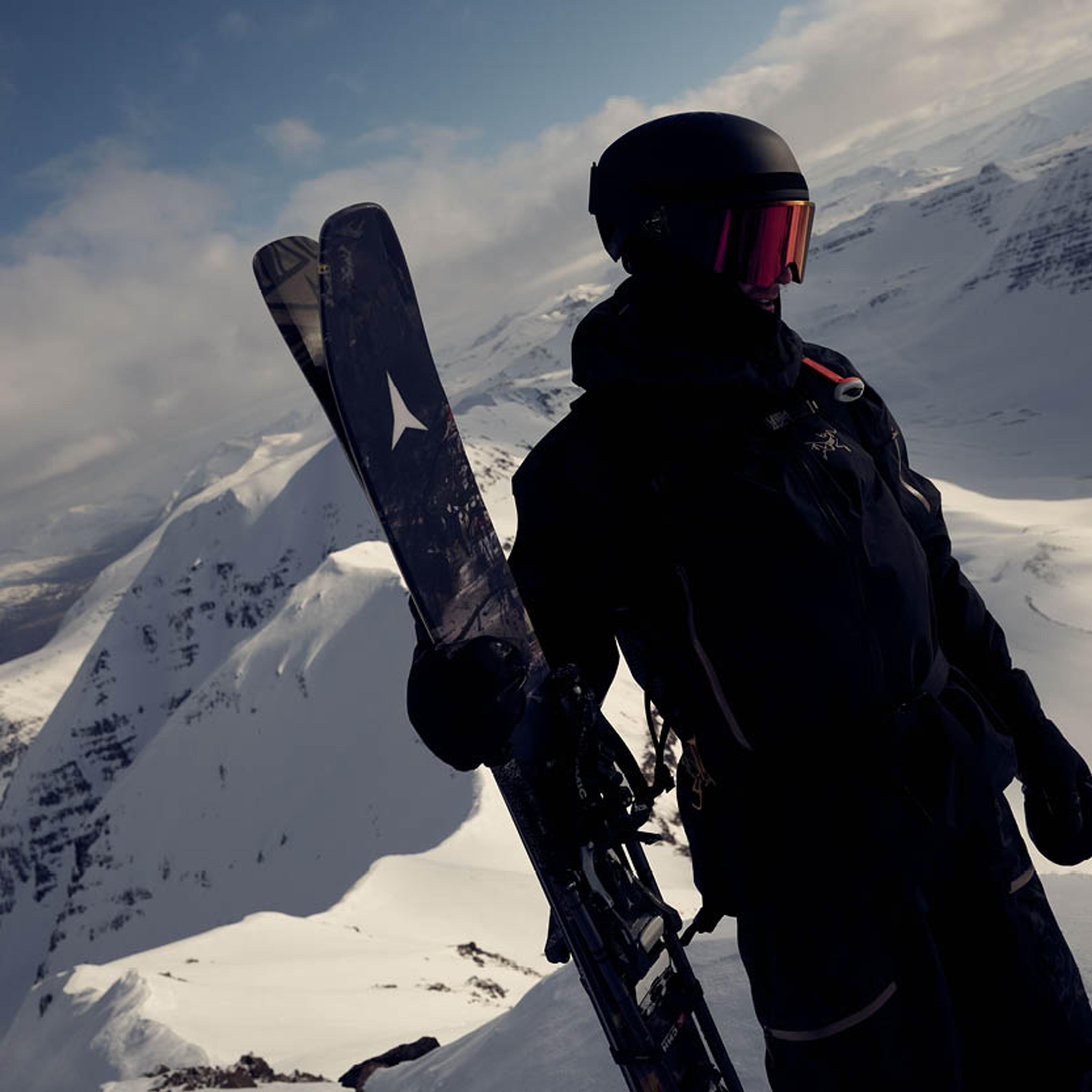 A skier wearing a black helmet and red goggles holds skis while overlooking a vast, snow-covered mountain range.