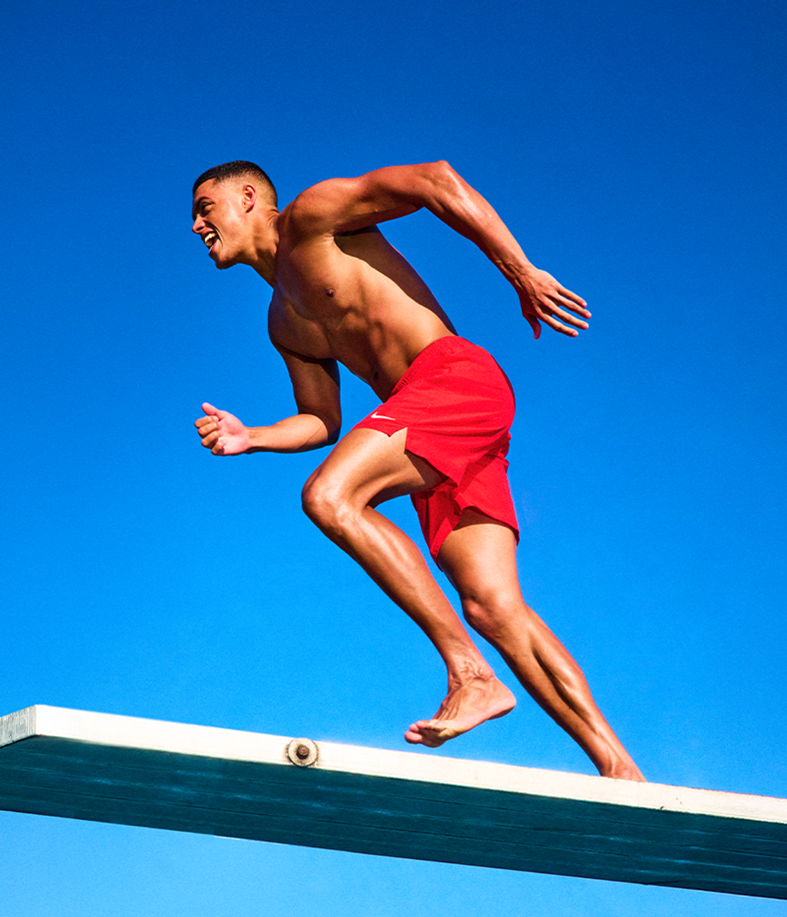 Shirtless, muscular man in red shorts runs barefoot on a diving board, mouth open in effort, against a blue sky.