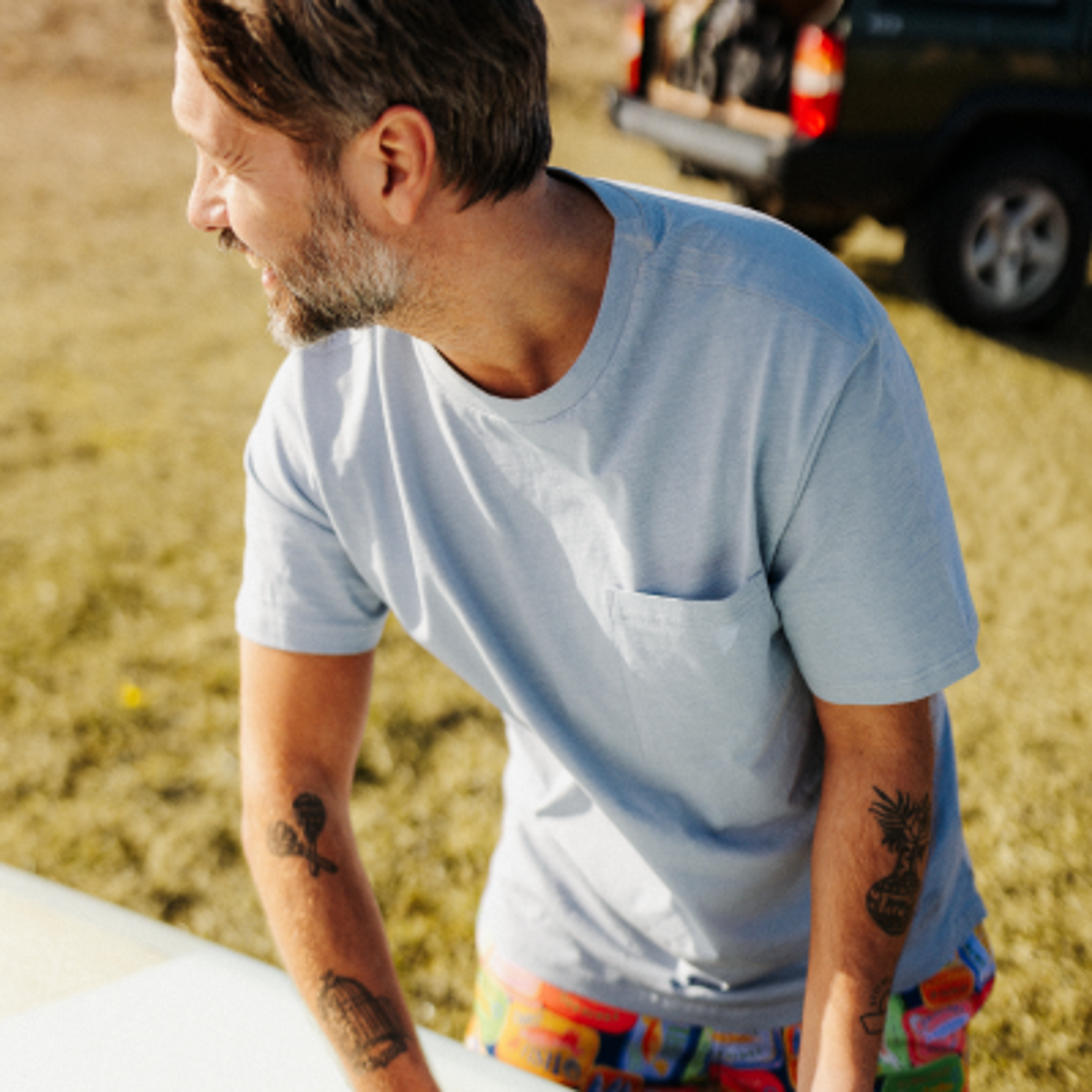 A smiling man with a beard and arm tattoos, wearing a light blue t-shirt and colorful shorts, prepares a surfboard outdoors.