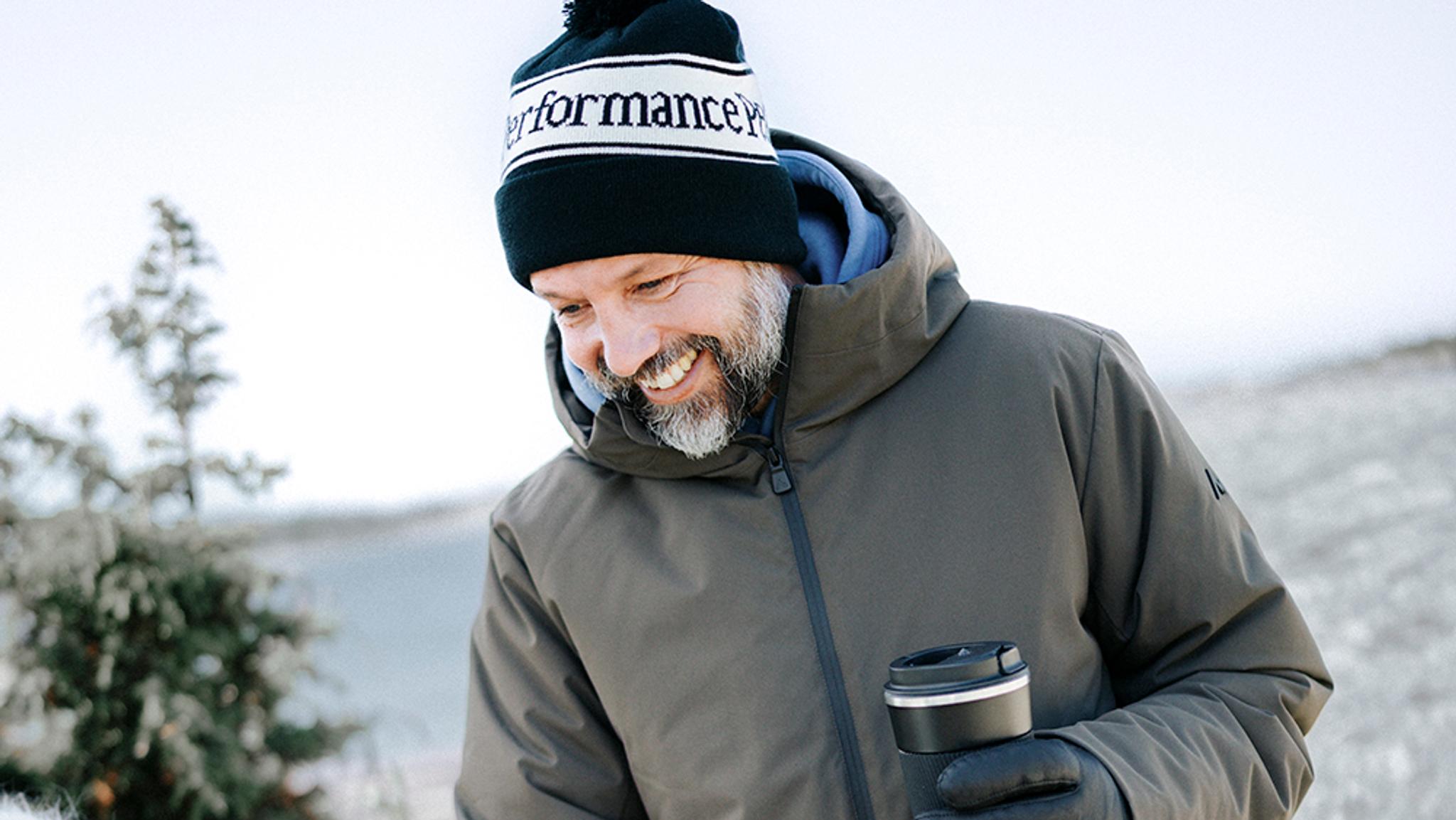 A smiling man in a winter jacket and beanie holds a travel mug in a snowy landscape.
