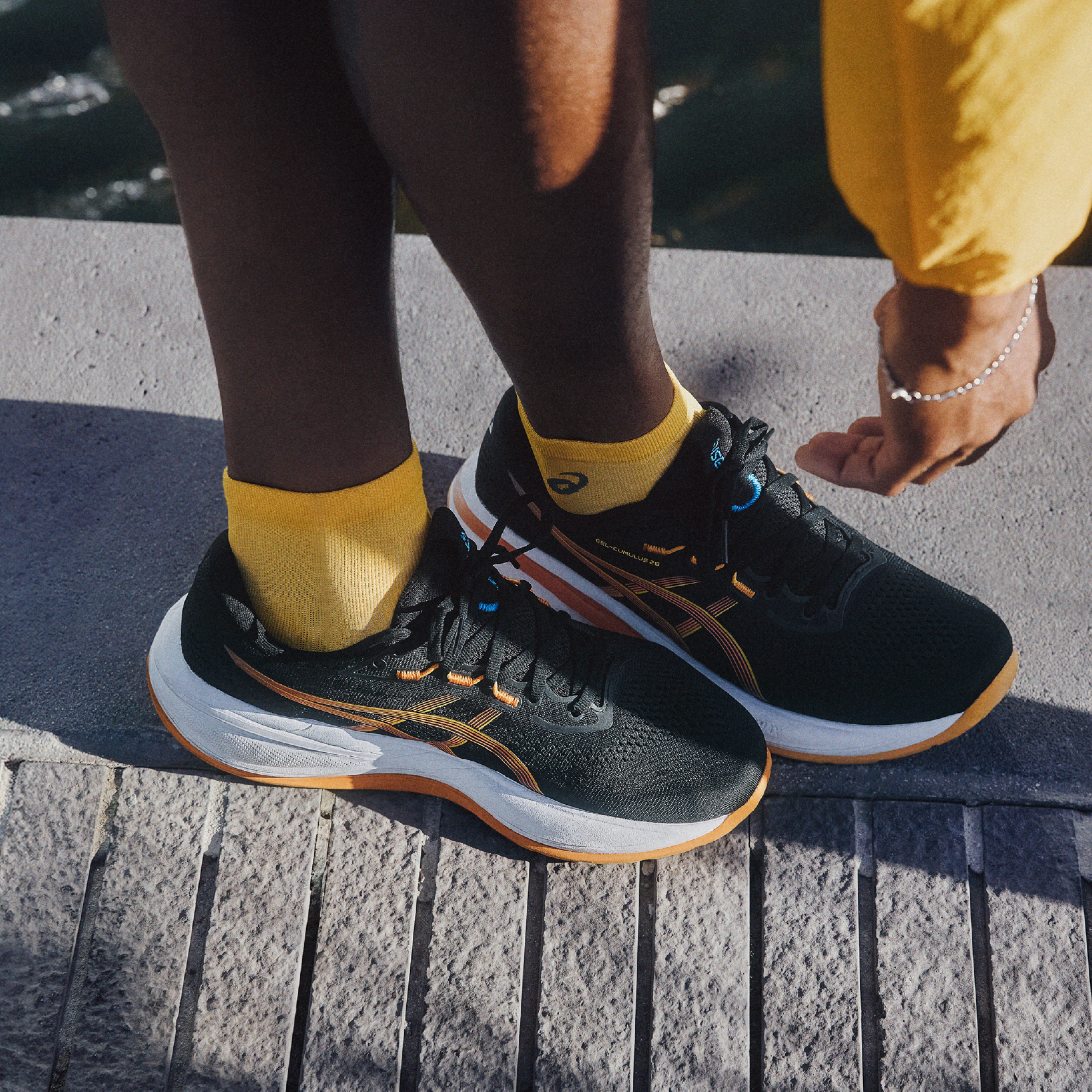 A person wears black Asics running shoes with orange accents and yellow socks on a stone surface.