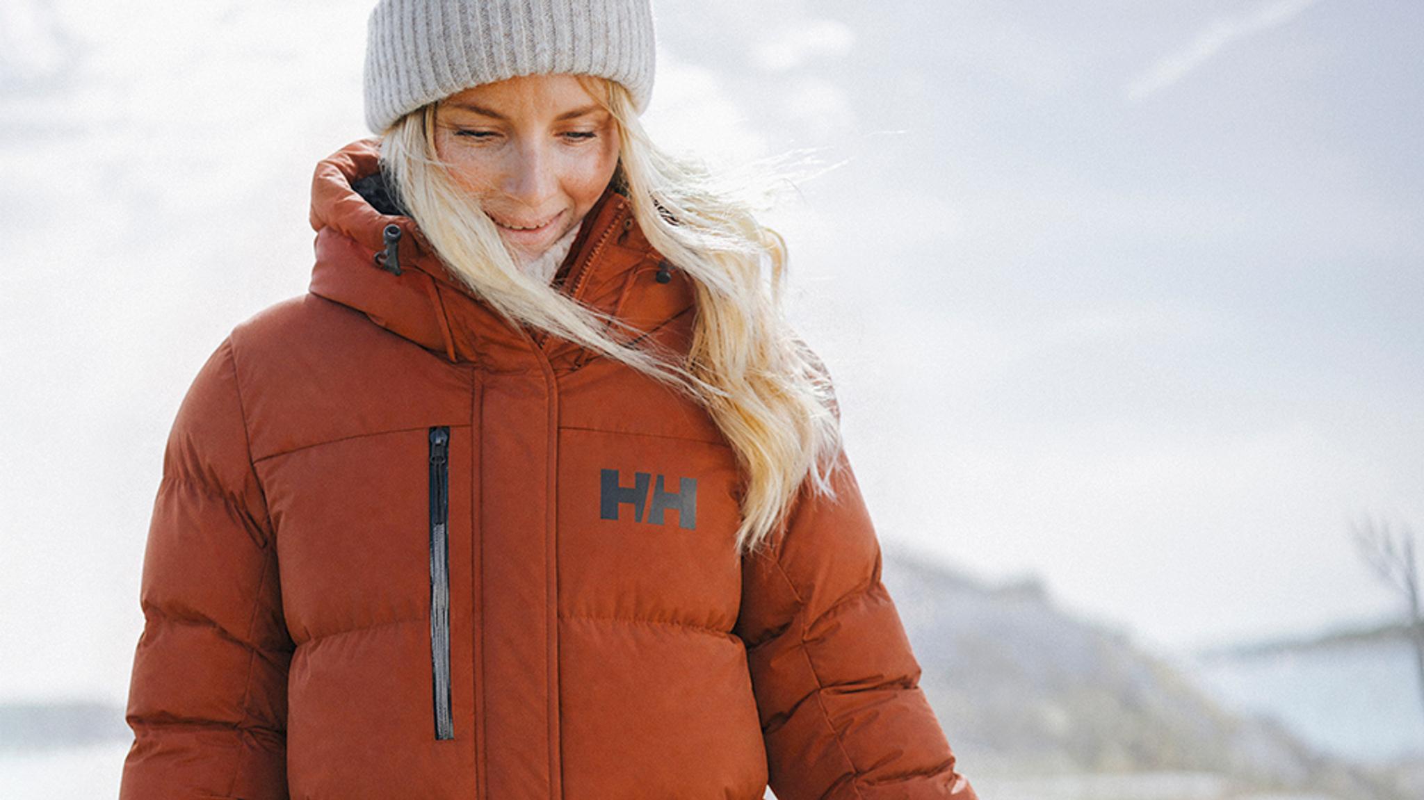 A smiling woman in a rust-colored Helly Hansen puffer jacket and a light beanie outdoors.