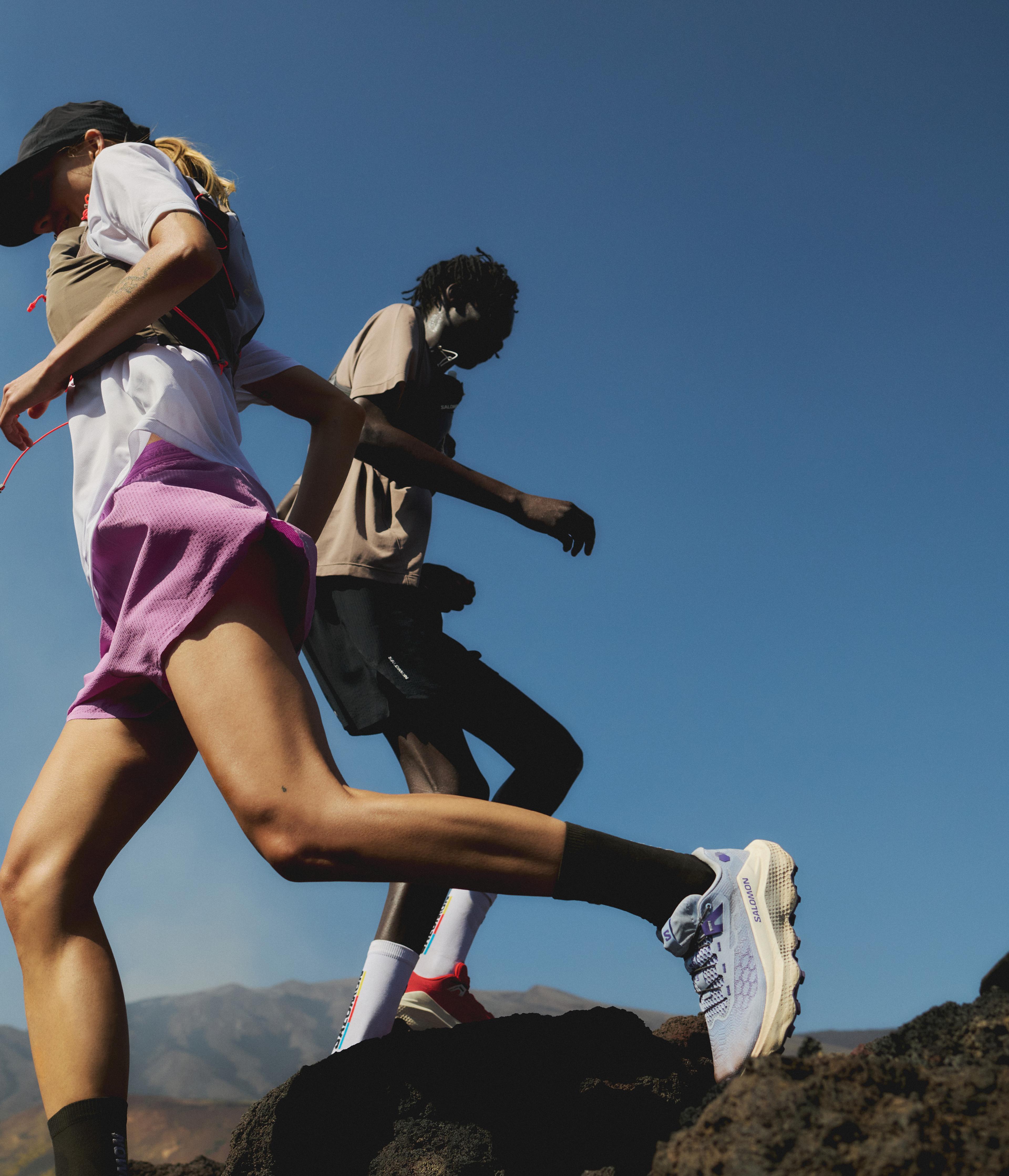 Two people trail running on rocky terrain under a clear blue sky.
