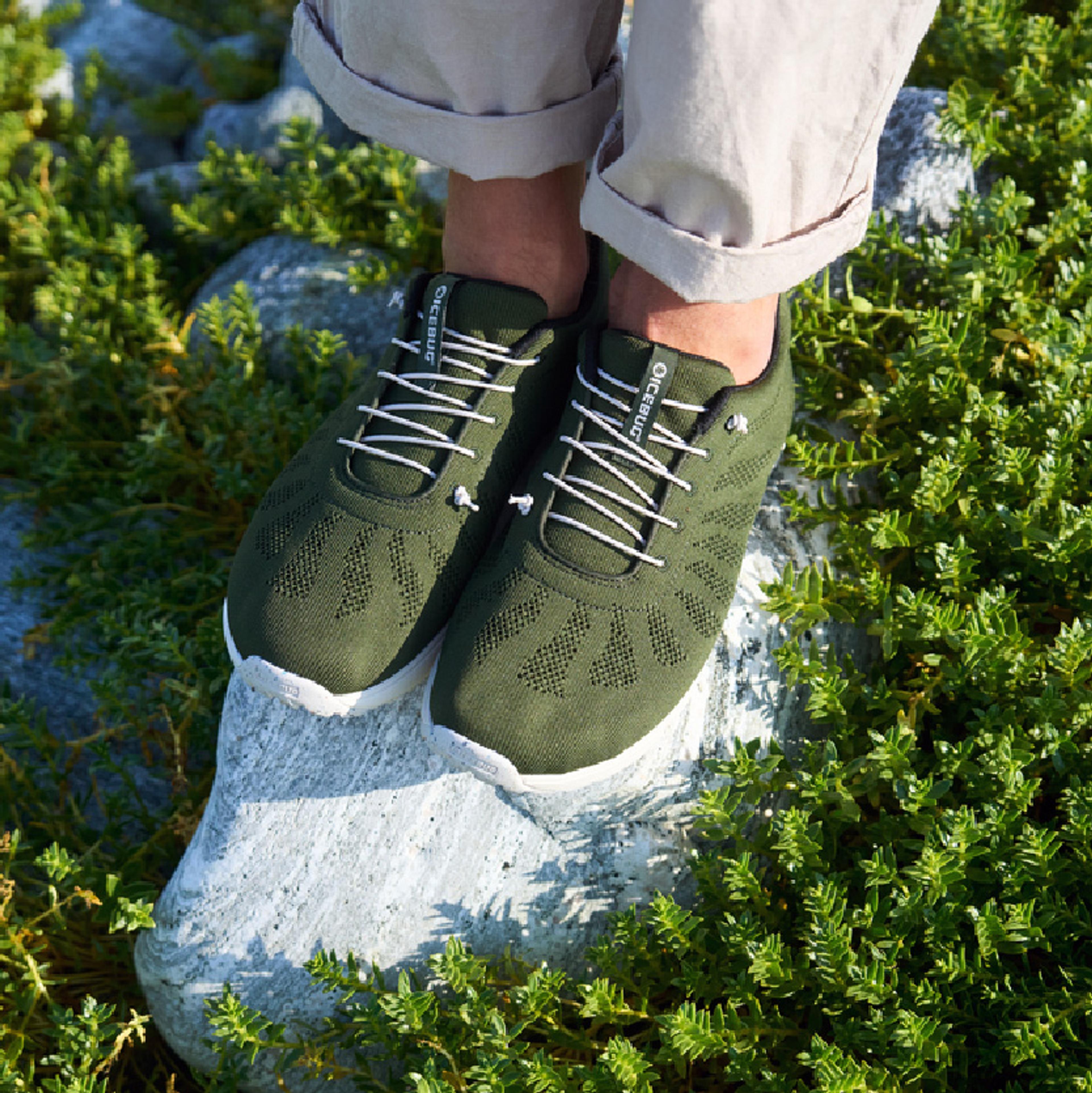 A pair of olive green sneakers with white laces and soles standing on a rock, surrounded by green foliage.