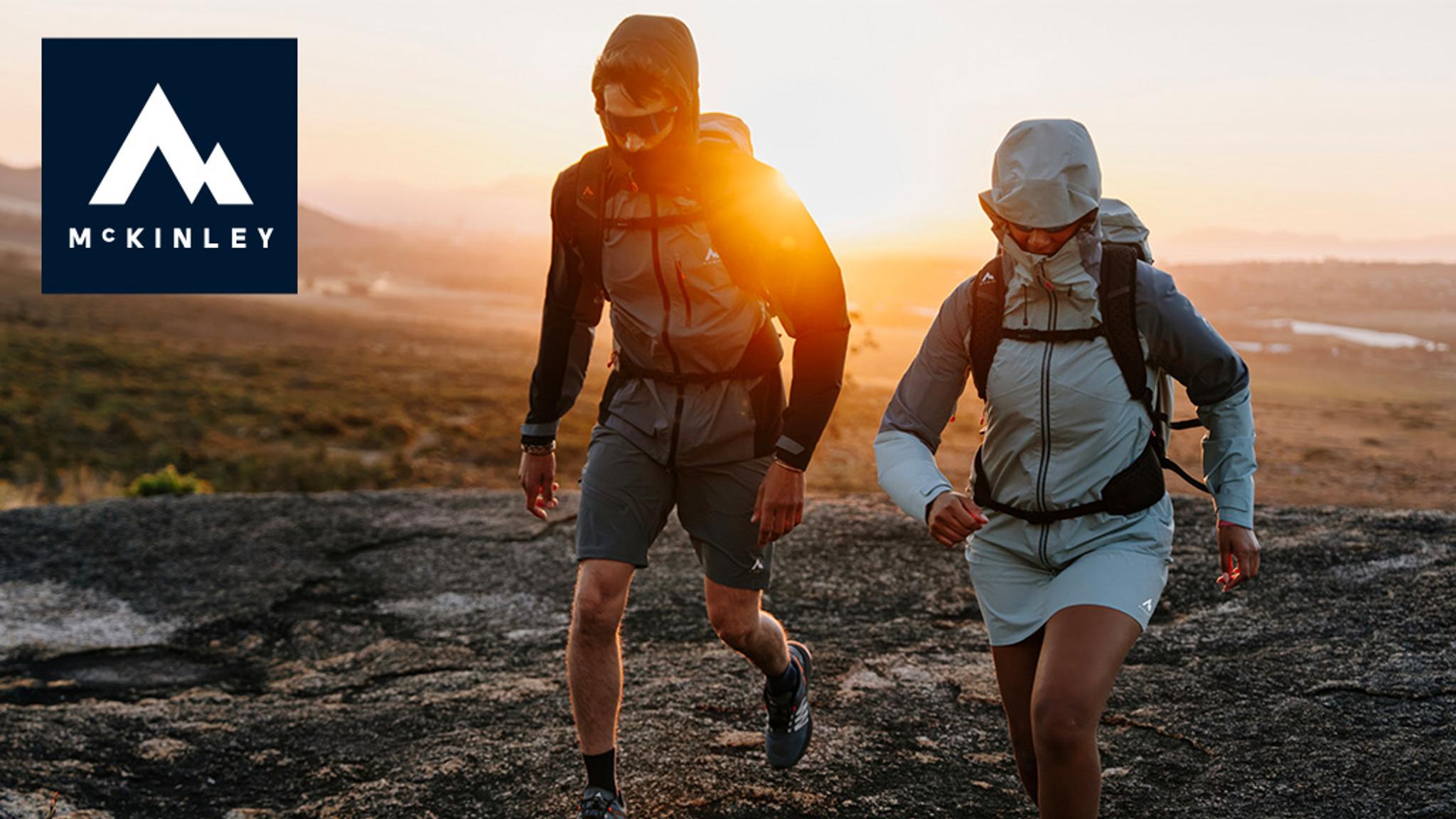 McKinley logo. Two hikers in outdoor gear ascend a rocky hill at sunset.