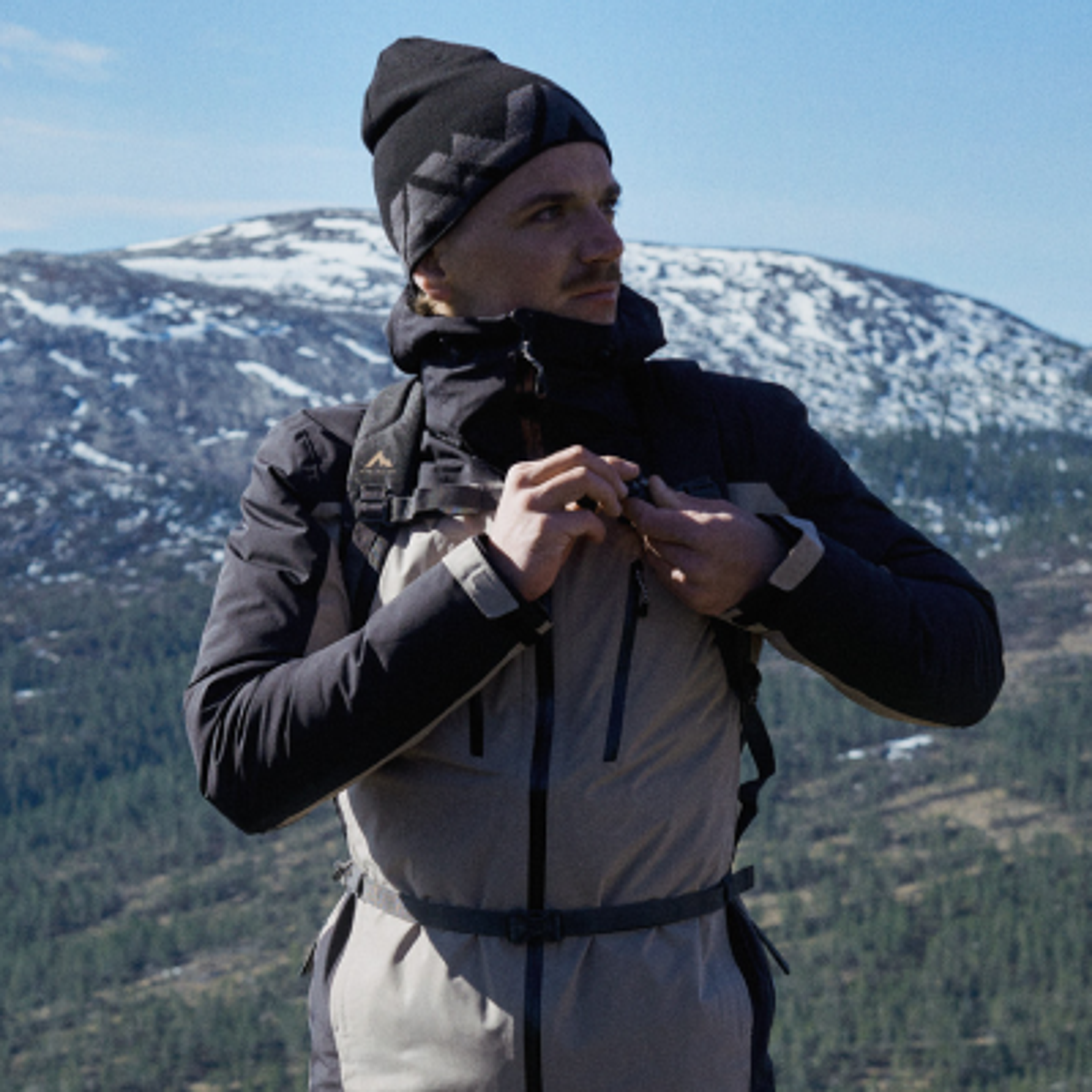 Man in a beanie and hiking jacket adjusting his zipper with a snowy mountain background.