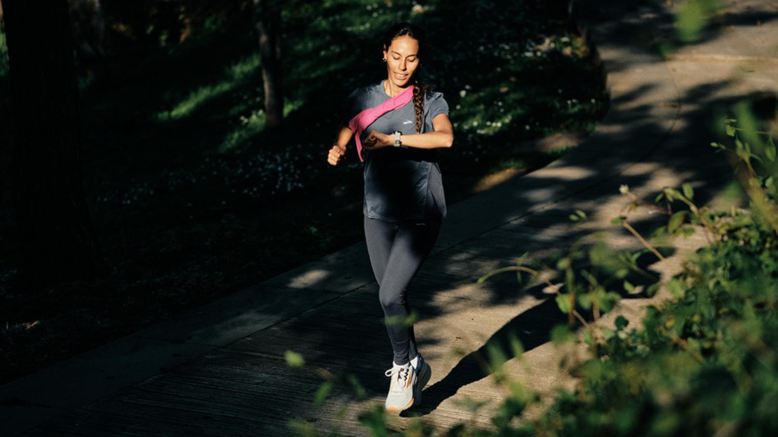 a woman is running down a path in the woods and looking at her watch .