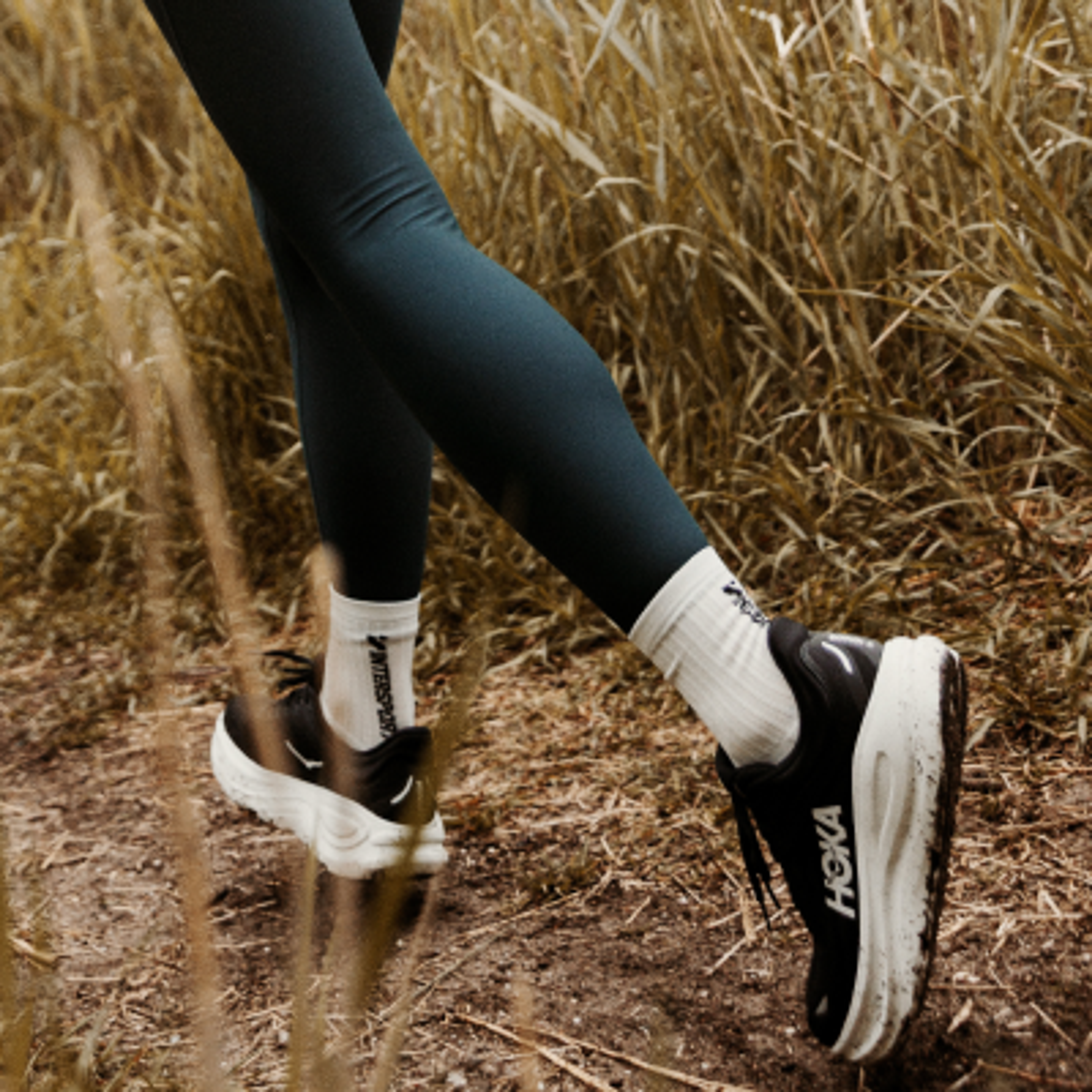 Legs in dark leggings, white Intersport socks, and black Hoka running shoes on a dirt trail lined with dry grass.
