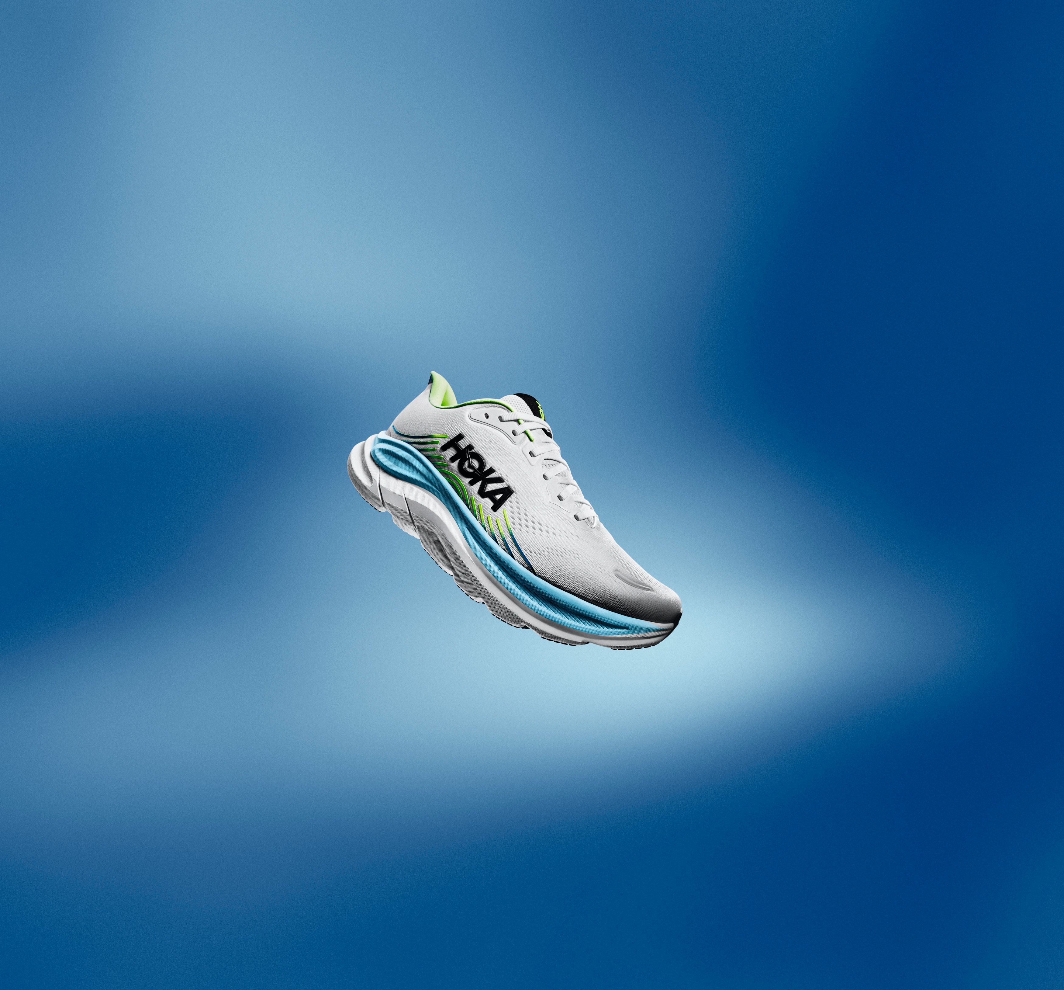 A white Hoka running shoe with blue and green accents floats against a blue background.