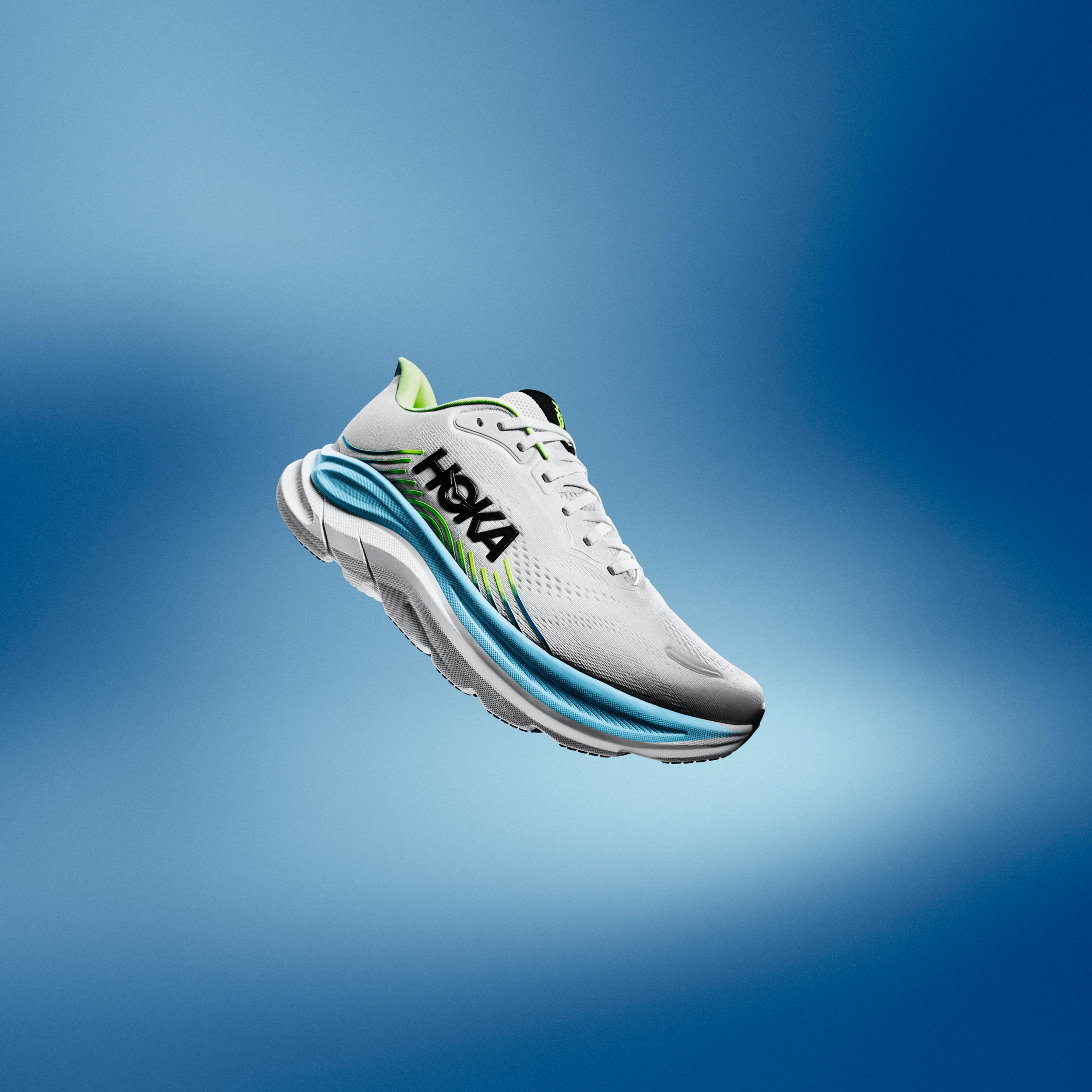 A white Hoka running shoe with blue and green accents floats against a blue background.