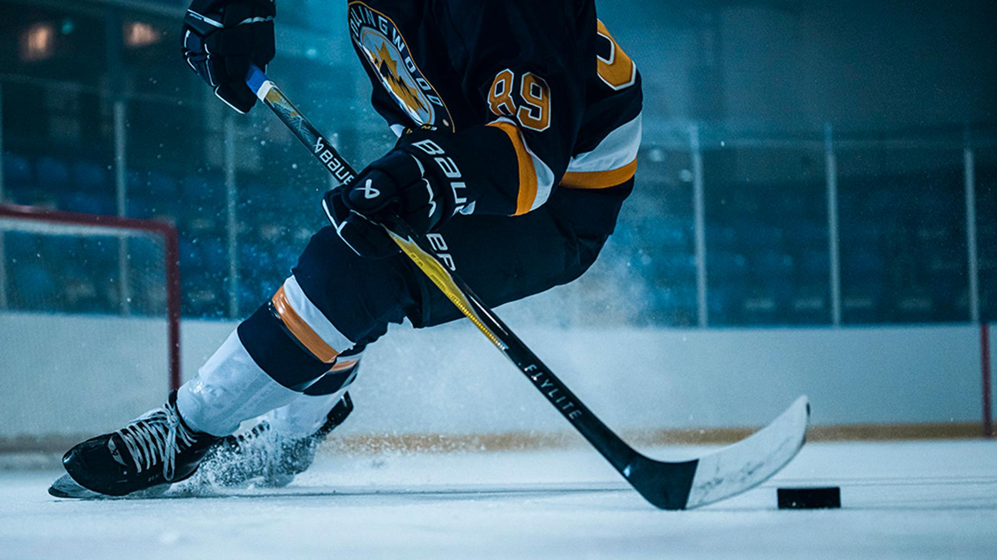 Hockey player in a dark and gold jersey skates on ice with a stick and puck, kicking up ice spray.