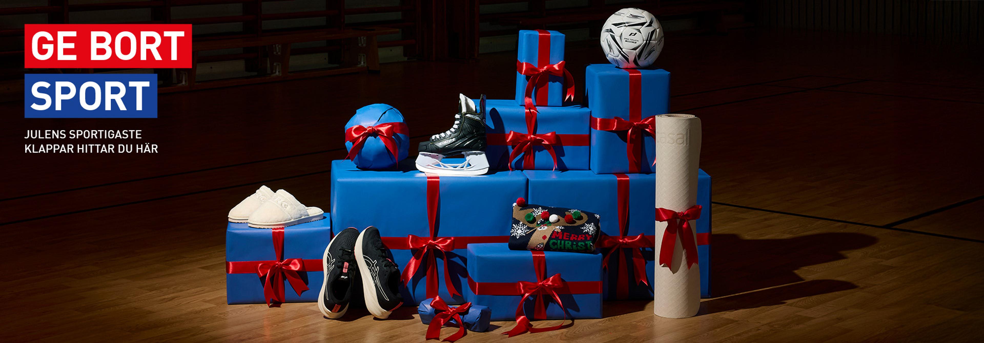 A collection of blue gift-wrapped boxes with red ribbons, sports gear including a football, ice skate, and sneakers, on a gym floor. Text: "GE BORT SPORT" (Give Away Sport), advertising Christmas gifts.