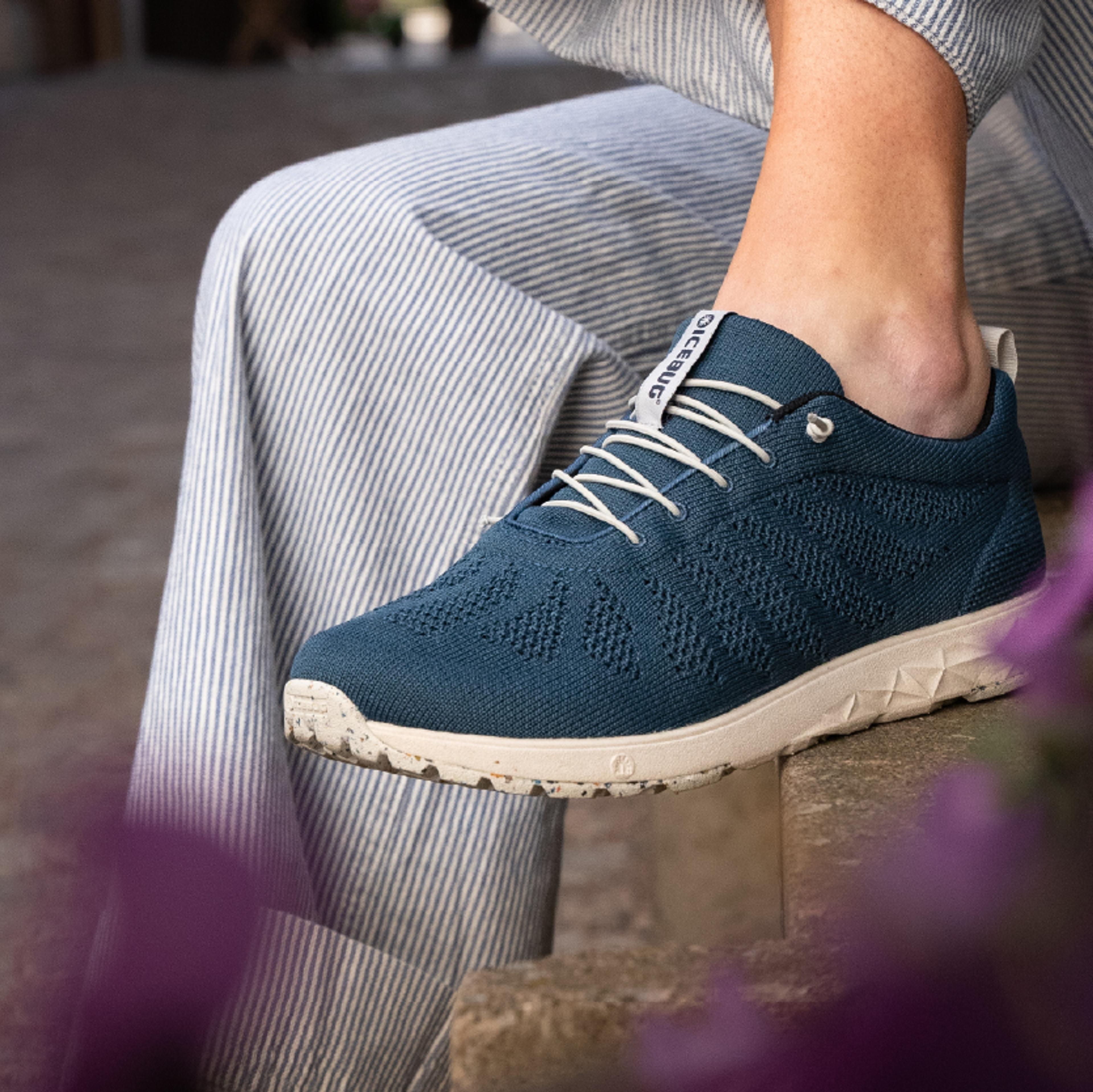 A person wears a blue knit Icebug sneaker with white laces, a speckled cream sole, and striped pants.