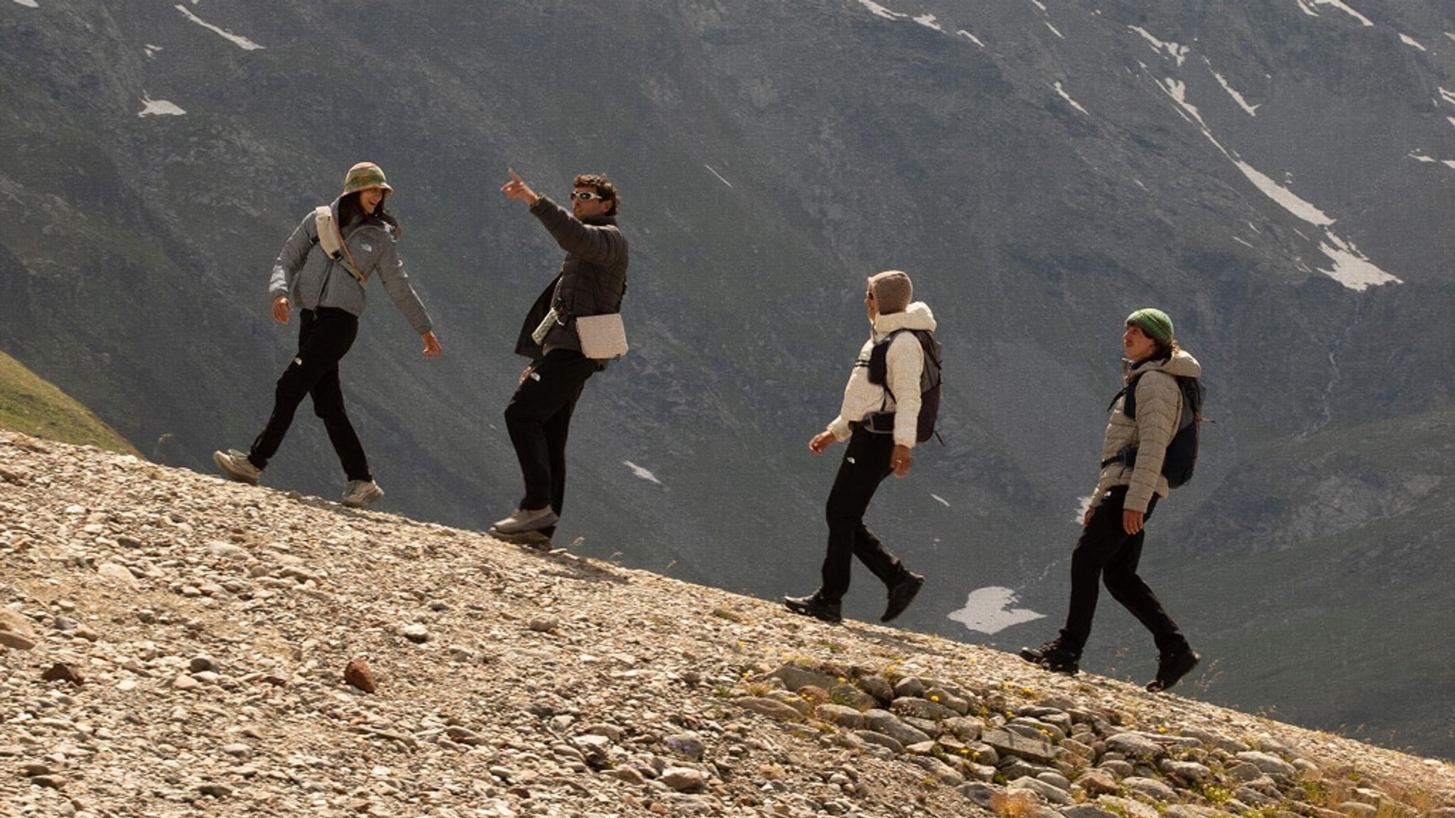 Four people hike uphill on a rocky mountain path, with one person pointing towards the distance.